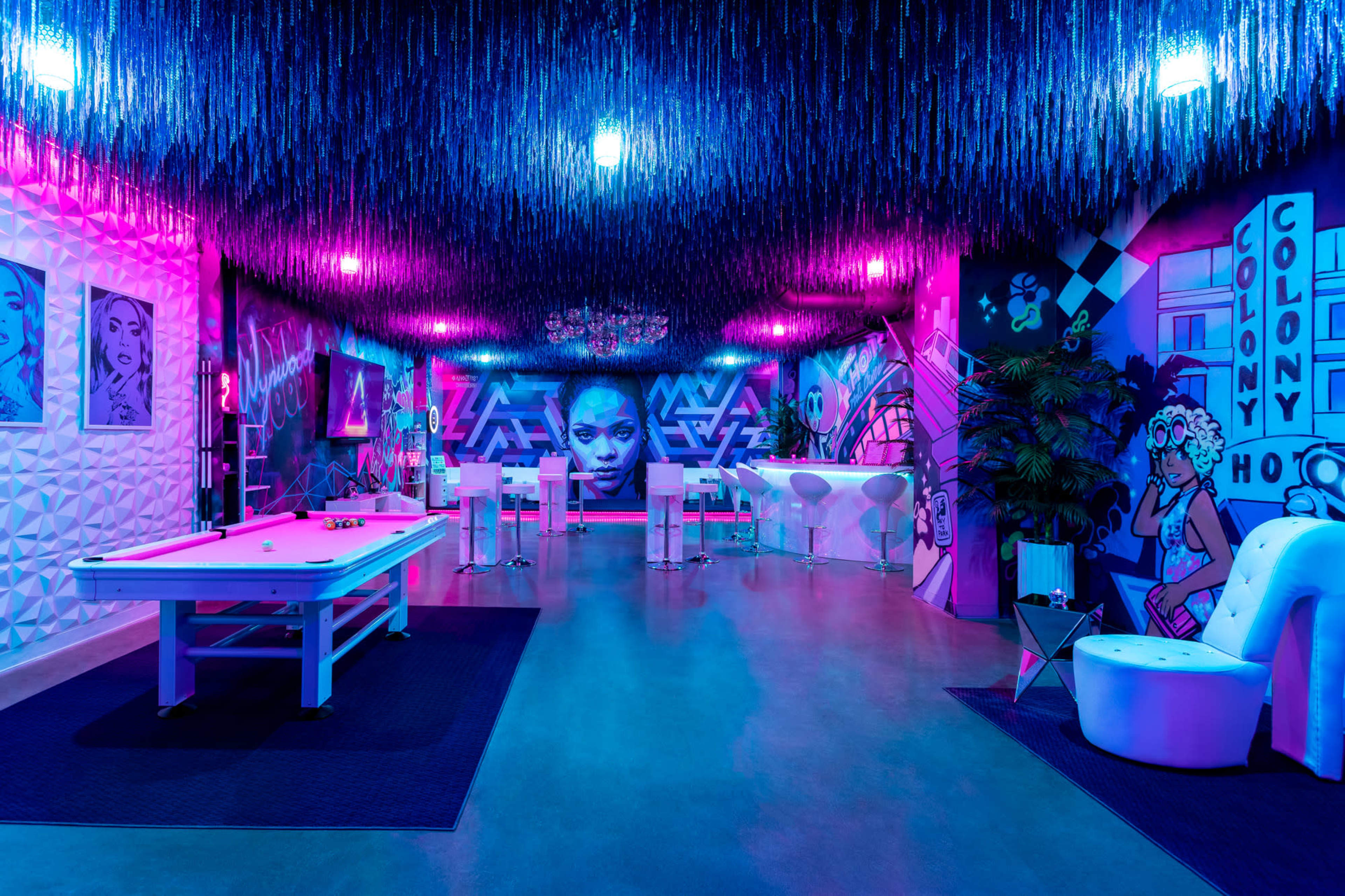 A vibrant lounge area with neon lights, colorful wall murals, and modern furniture, featuring a pool table in the center.