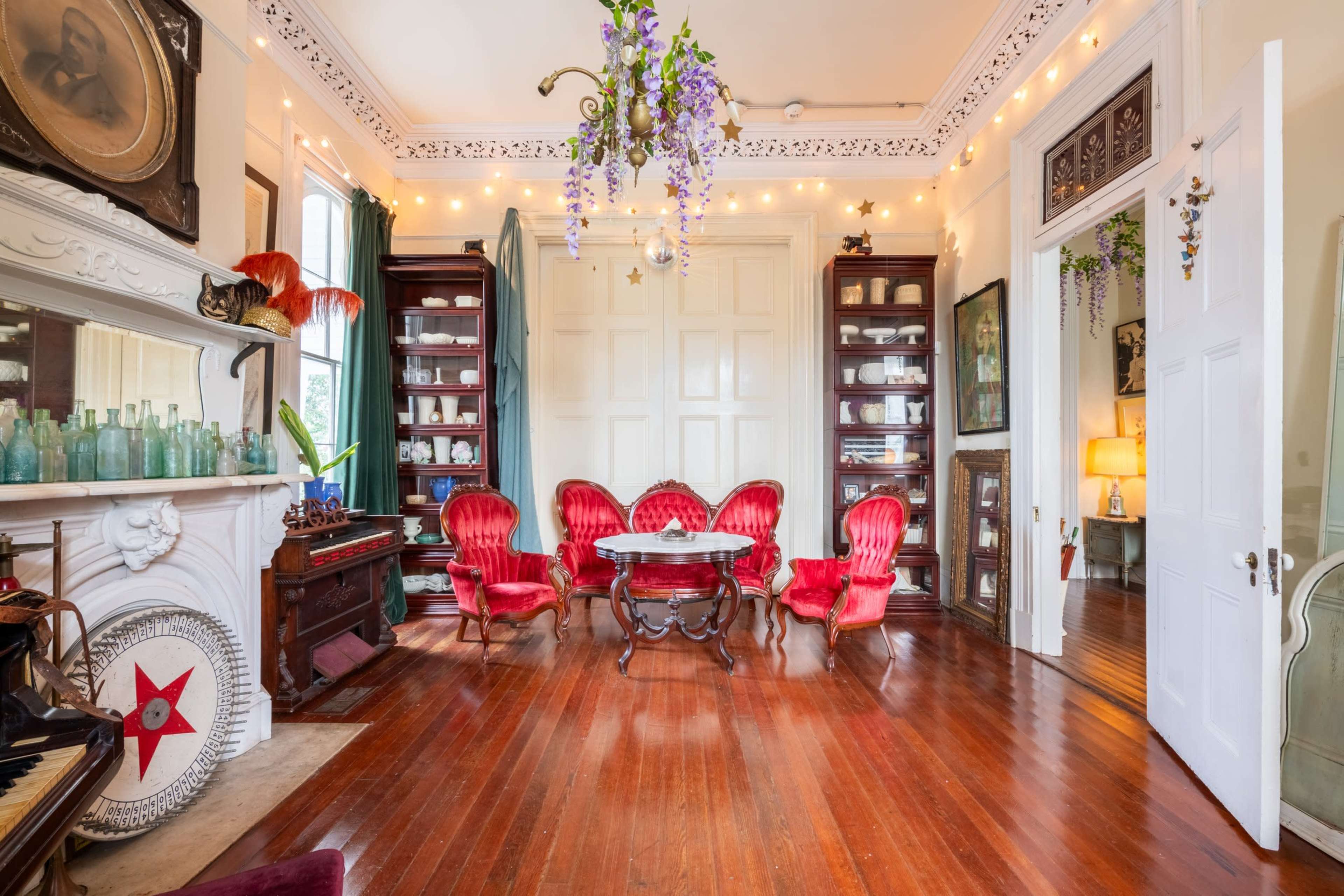 A cozy room features red velvet chairs surrounding a central table, with wooden floors, a fireplace, and shelves filled with collectibles.