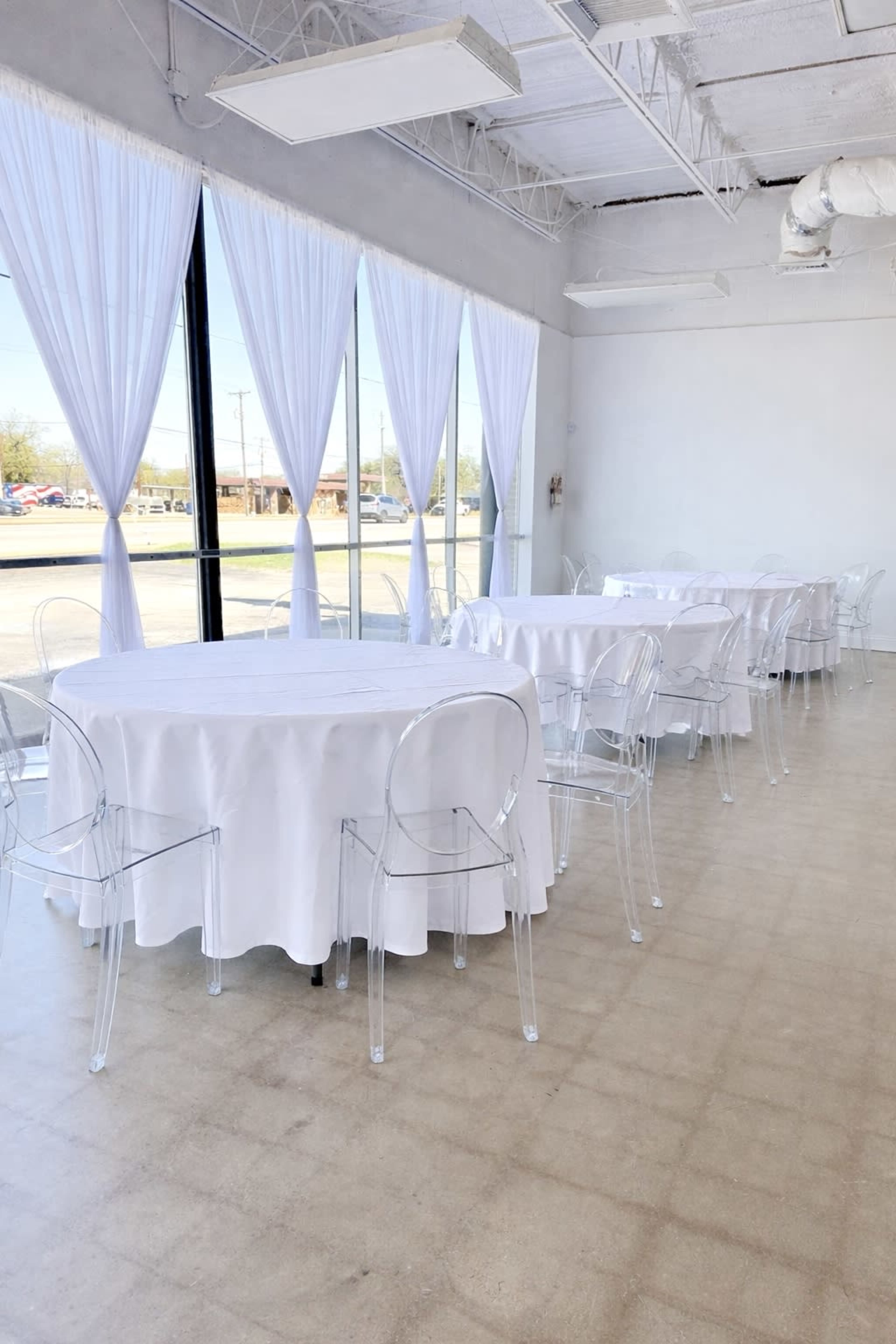 The image shows a spacious room with several round tables covered in white tablecloths and surrounded by clear ghost chairs, with large windows allowing natural light to enter.