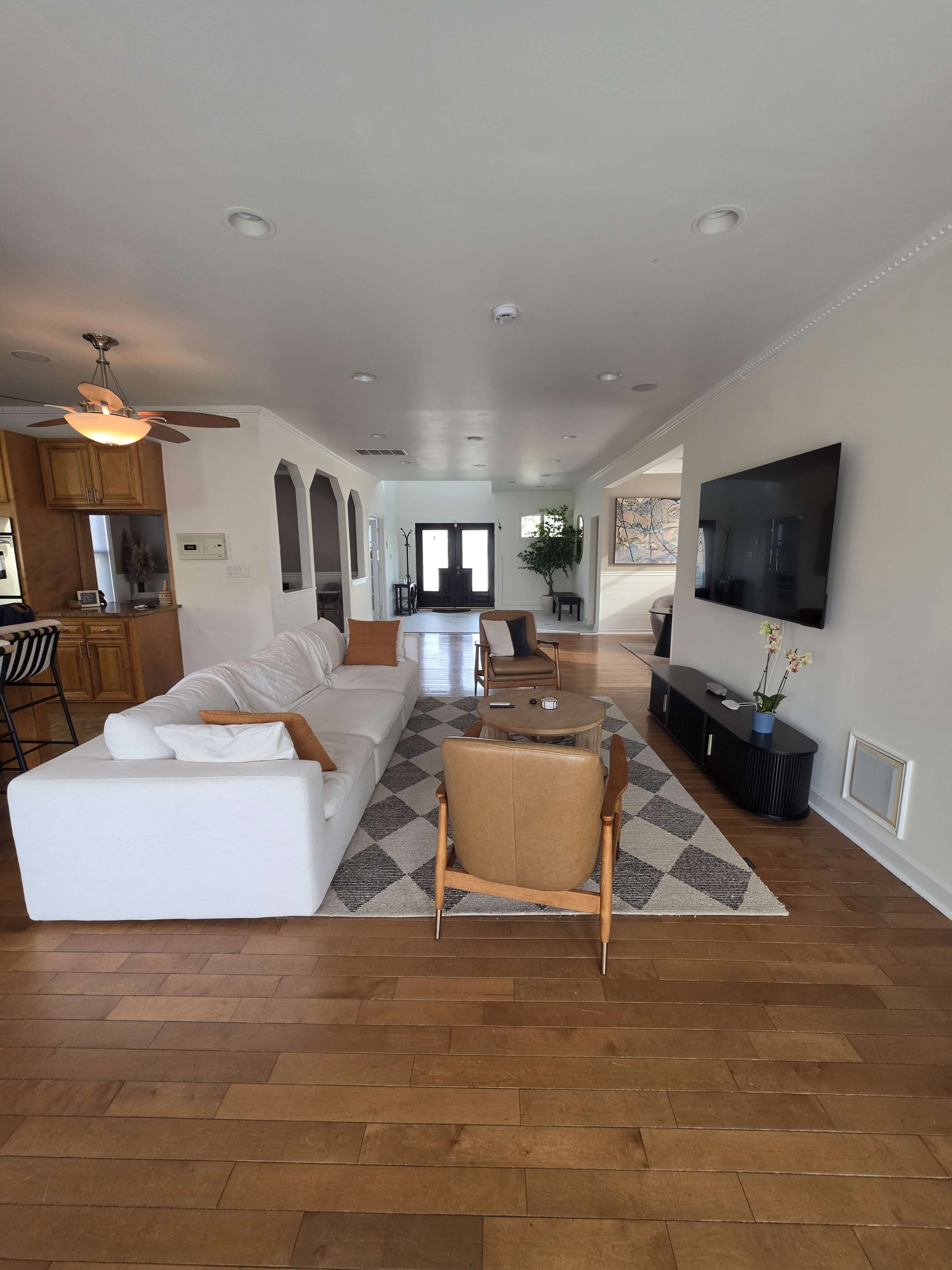 The image shows a spacious living room featuring a white sofa, two brown chairs, and a wall-mounted television, with wooden flooring and natural light coming through large windows.