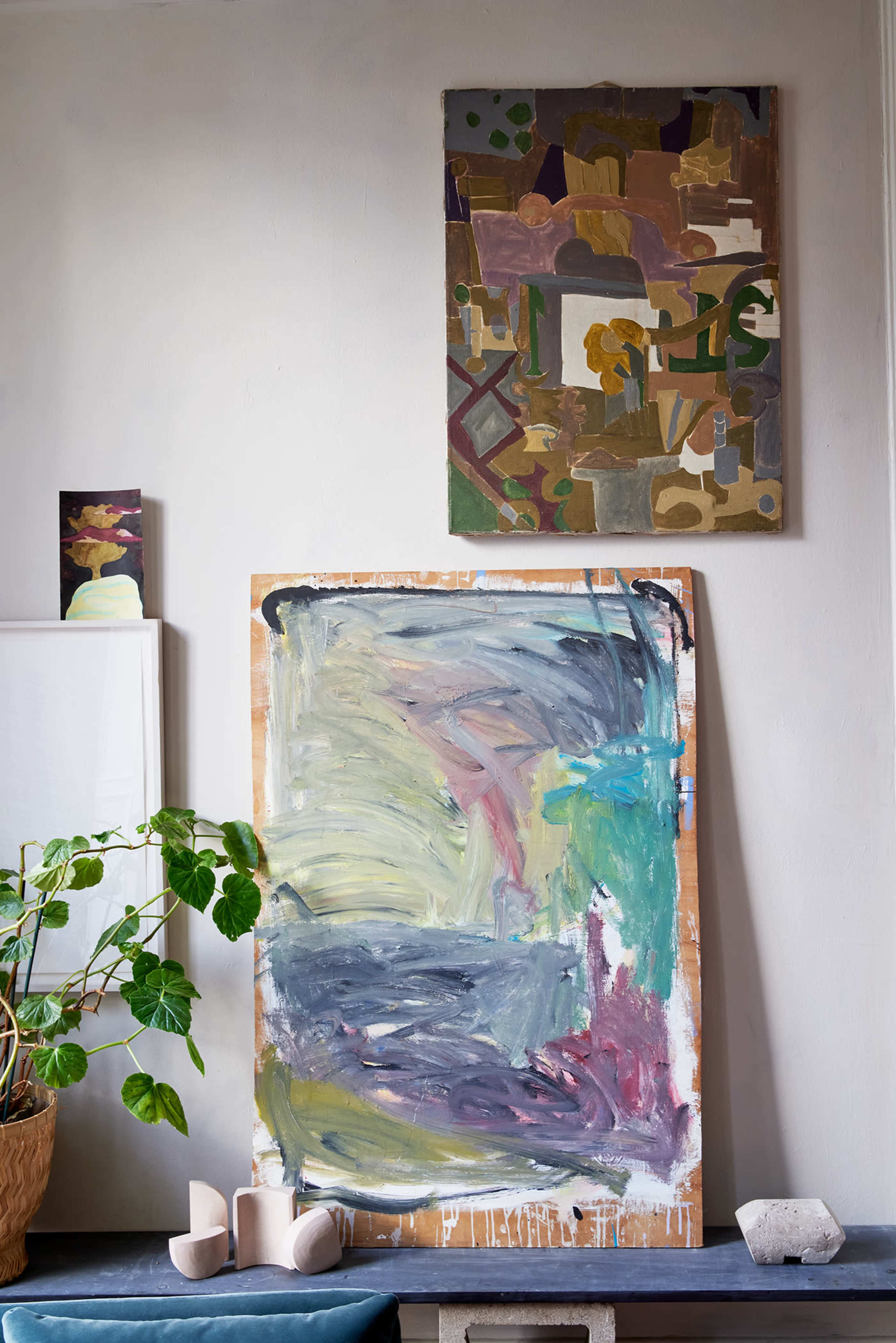 A collection of abstract paintings is displayed on a wall, with a vibrant, large canvas in the foreground and smaller artworks above it, alongside a potted plant and a few decorative objects on a shelf.