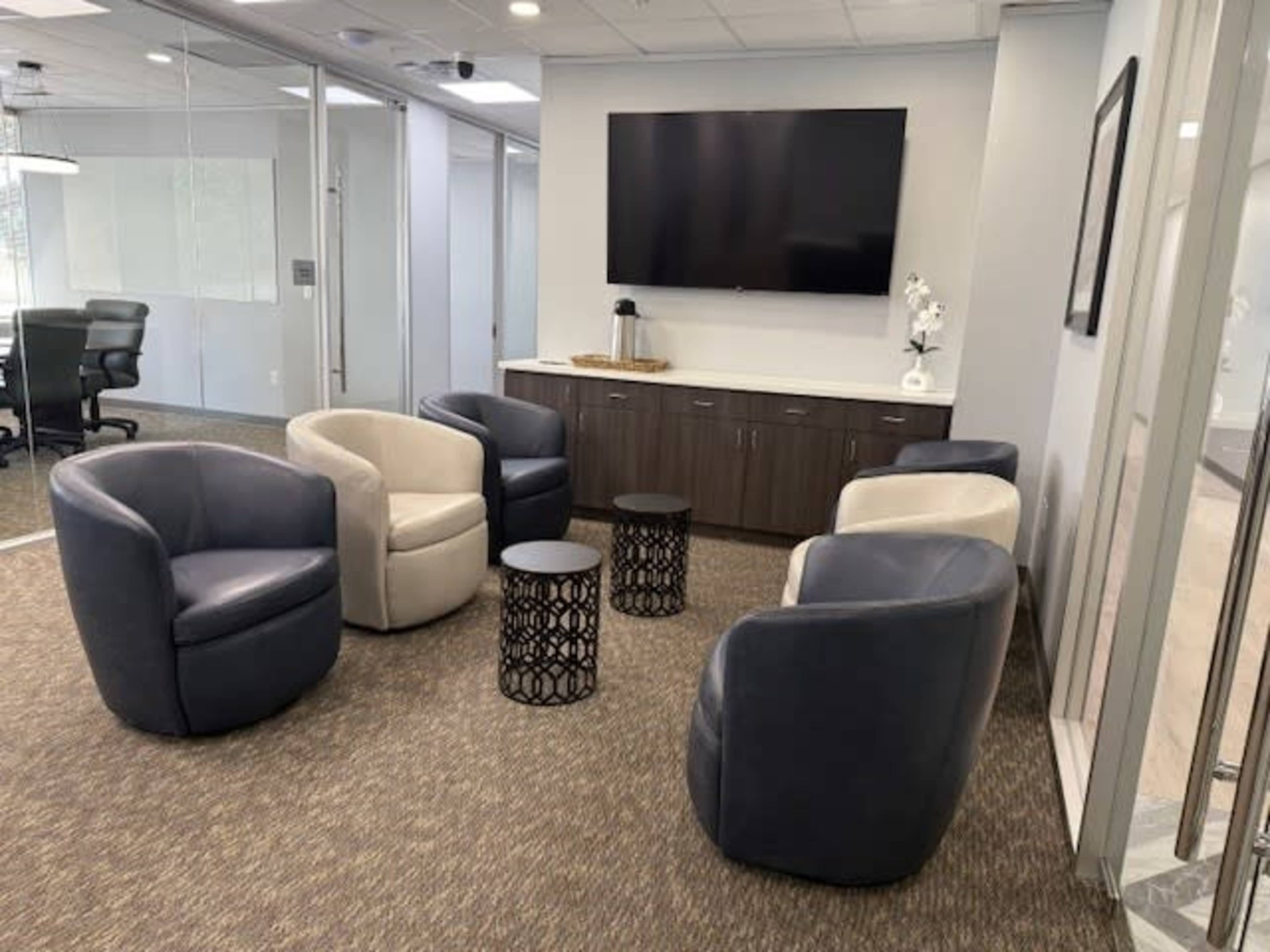 The image shows a modern office waiting area with four upholstered chairs arranged around two small side tables and a wall-mounted television.
