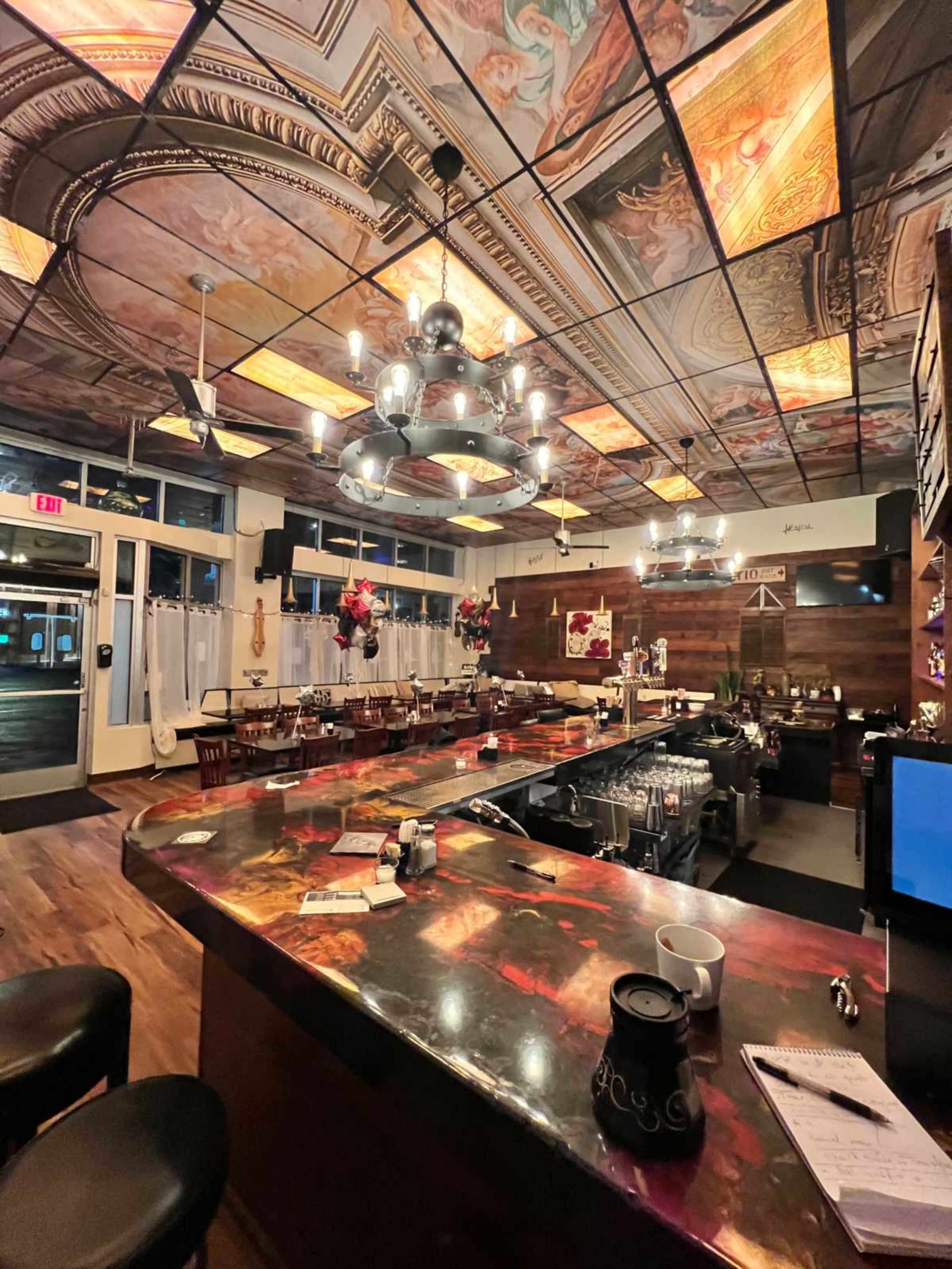 The image shows a restaurant interior featuring ornate ceiling artwork, a long bar with stools, and tables arranged in a warm, inviting atmosphere.