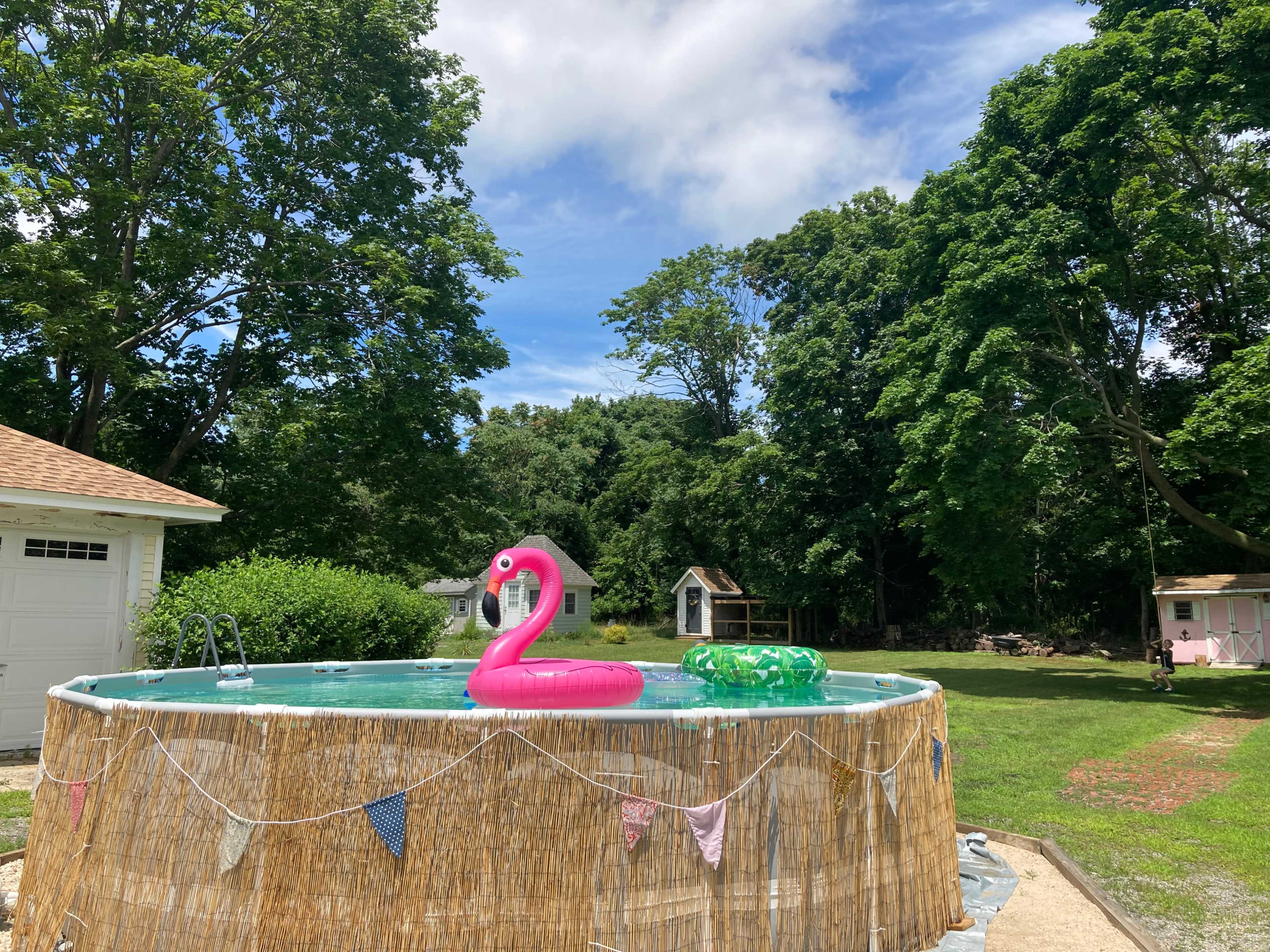 The image shows a backyard with a round above-ground pool, featuring a pink flamingo float and surrounded by lush trees and a grassy area.