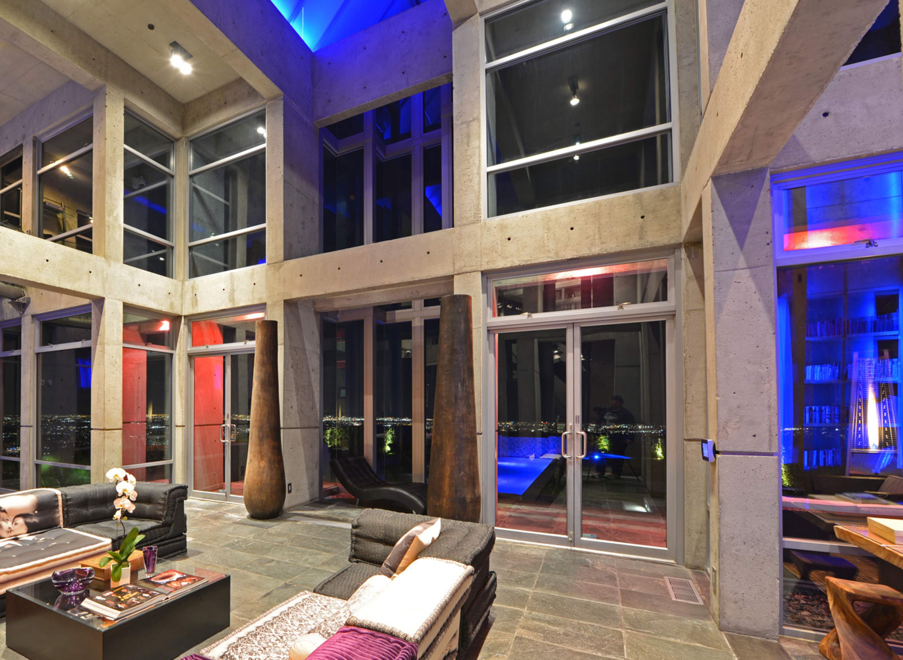 The interior of a modern, open-concept living space features large glass windows, concrete walls, and minimalistic furniture arranged around a stone floor, with views of city lights beyond.