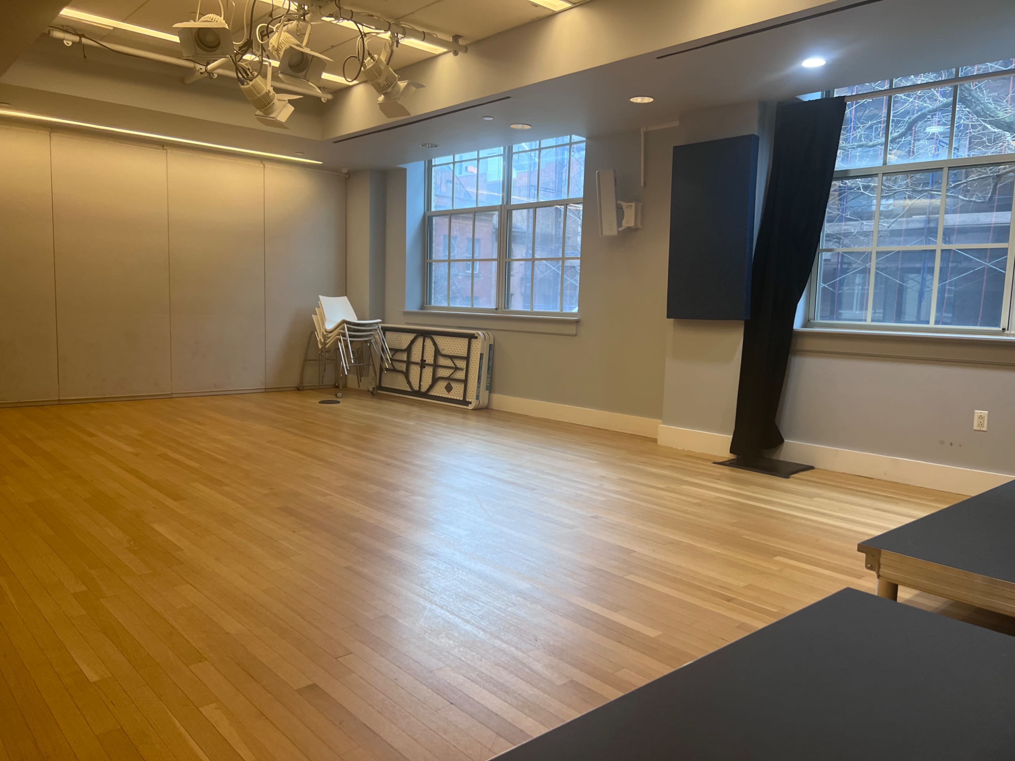 The image shows a spacious, empty room with hardwood flooring, large windows, and a few stacked chairs against the wall.