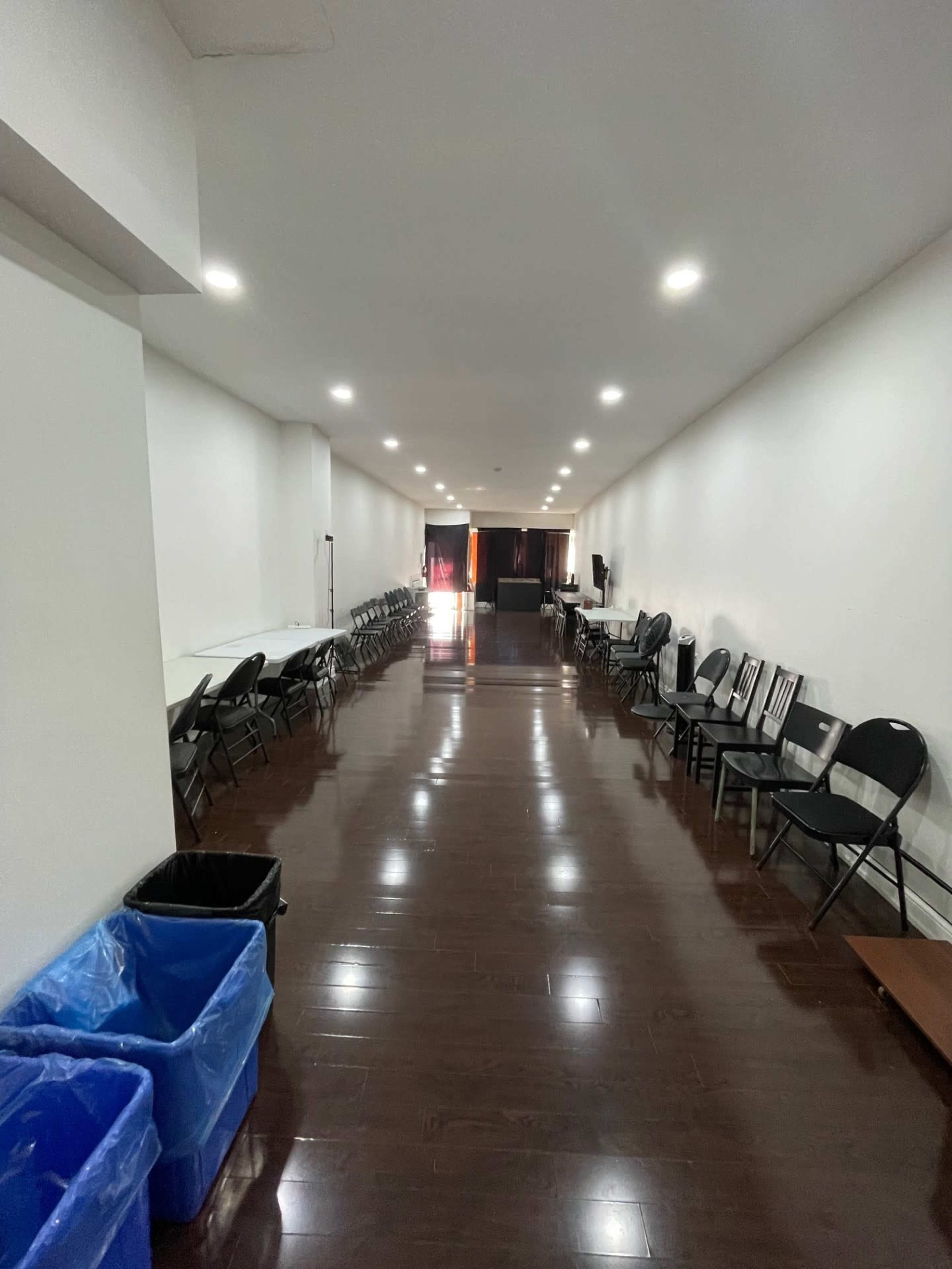 The image shows a long corridor lined with chairs on both sides and several tables against the walls, with two blue trash bins at one end.