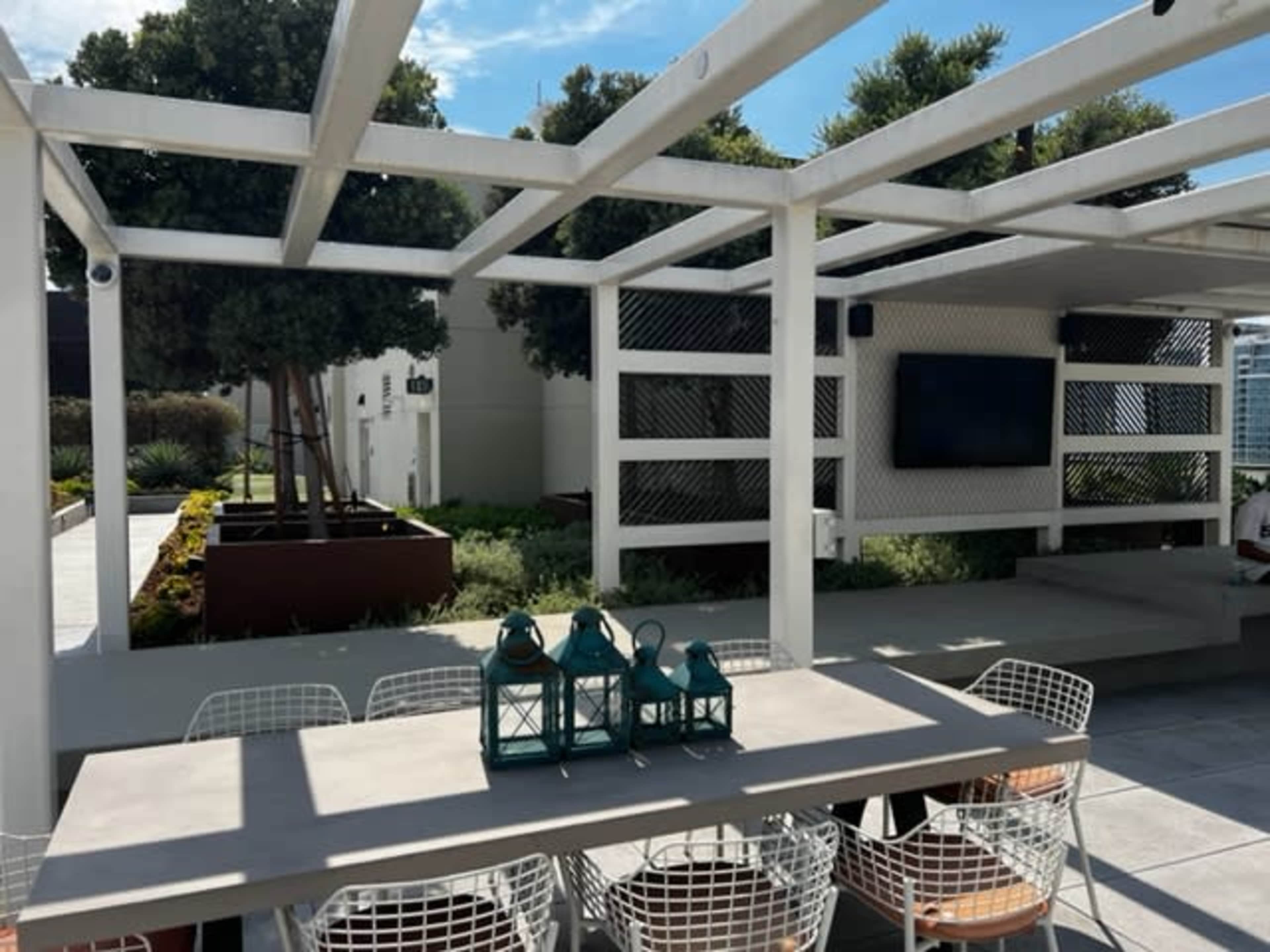 An outdoor seating area with a large table, lanterns, and a television mounted on a wall, surrounded by landscaping and trees.