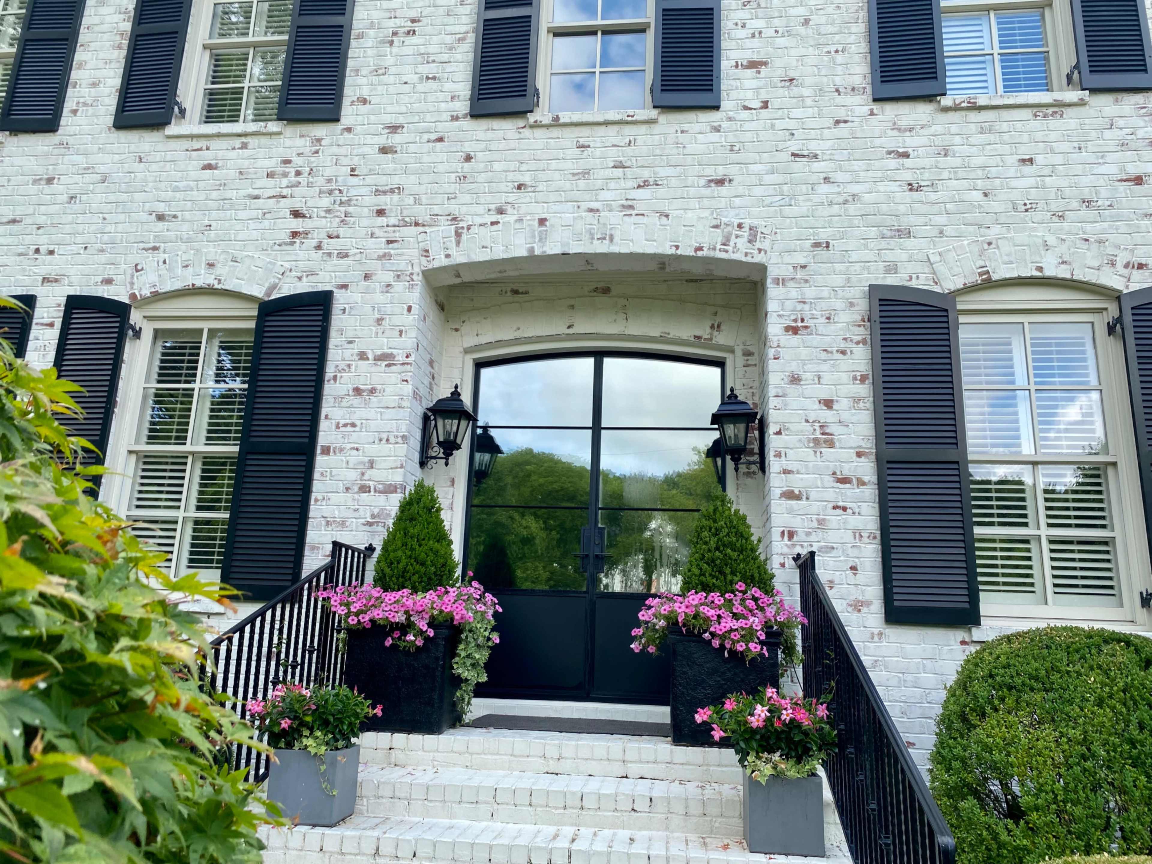 The entrance of a white brick house features large black shutters, a glass door, and planters with pink flowers on either side of the staircase.