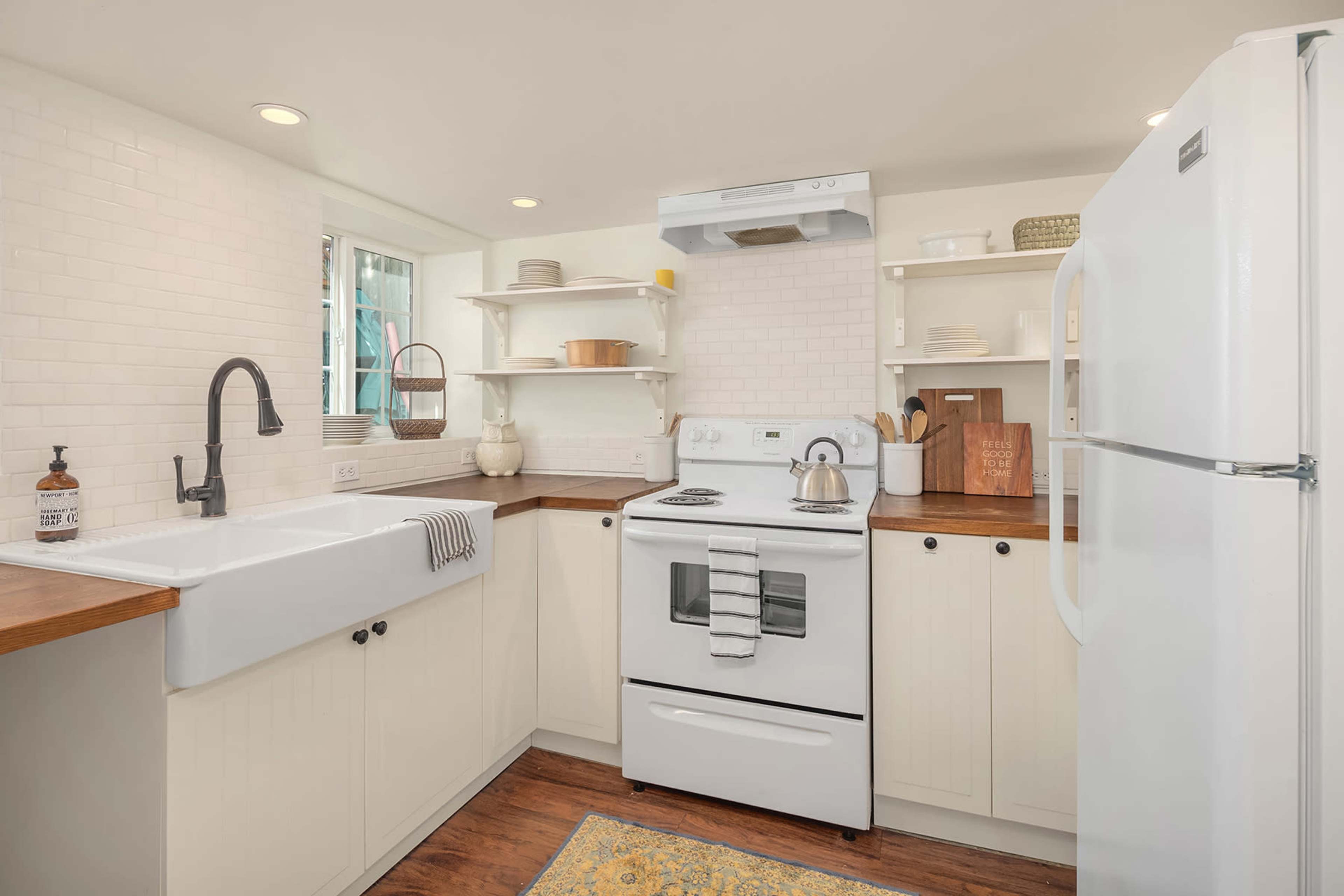 The image shows a compact kitchen with a farmhouse sink, white cabinetry, a stove, and open shelves displaying dishes.