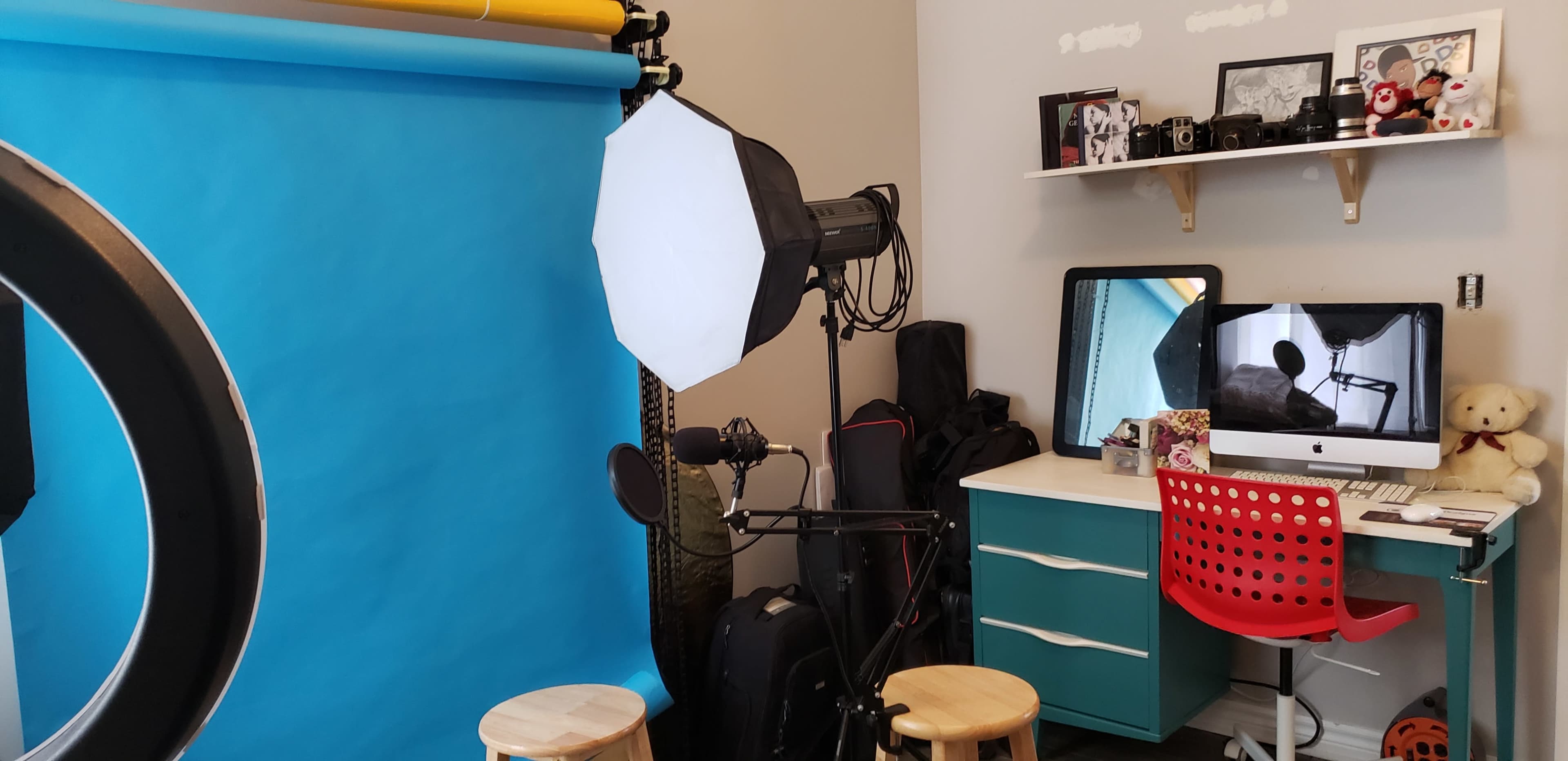 The image shows a photography setup with a blue backdrop, lighting equipment, a desk with a computer, and a red chair.