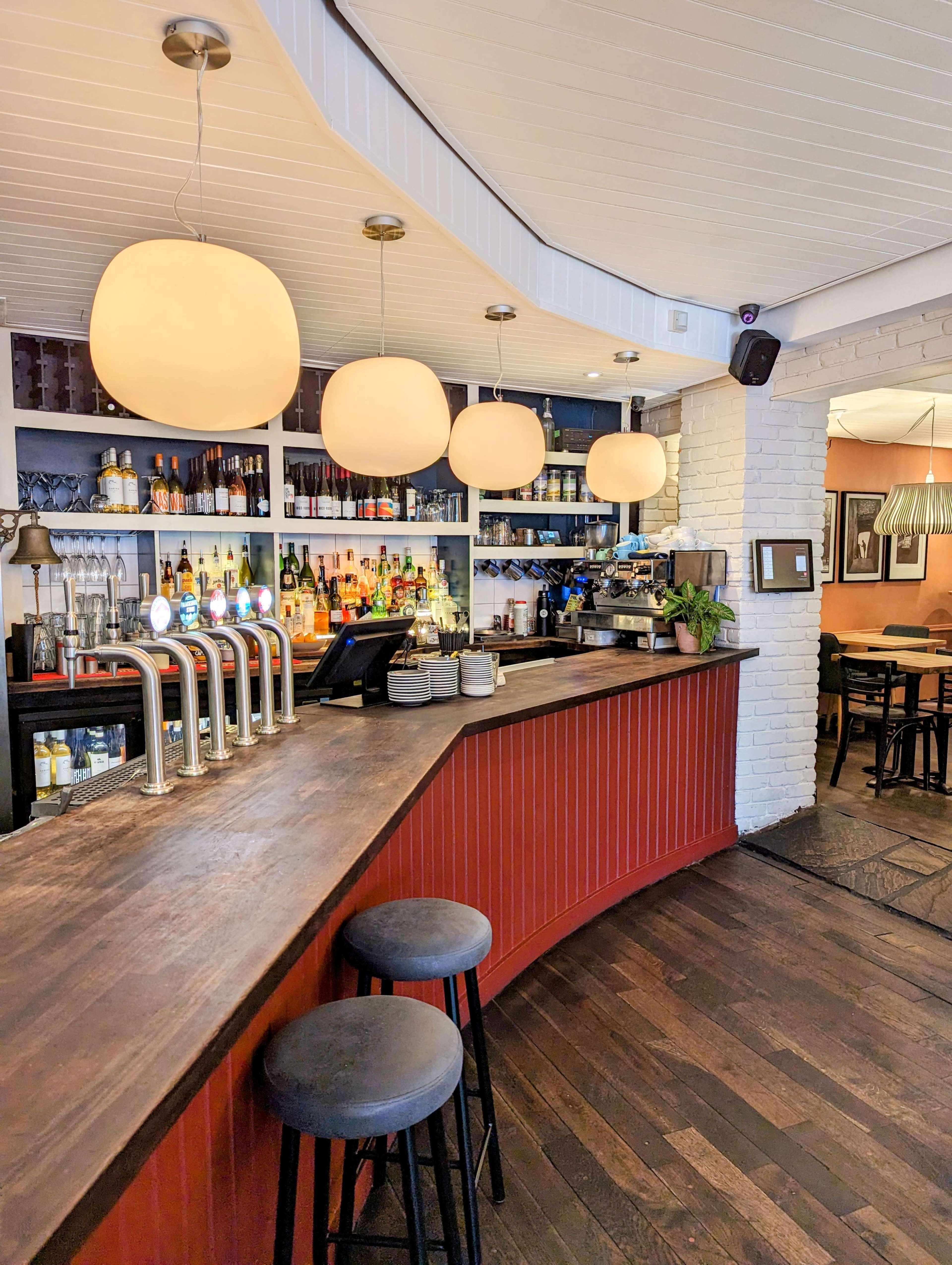 A bar area features a curved wooden counter, three black stools, and pendant lighting above, with shelves displaying various bottles behind the bar.