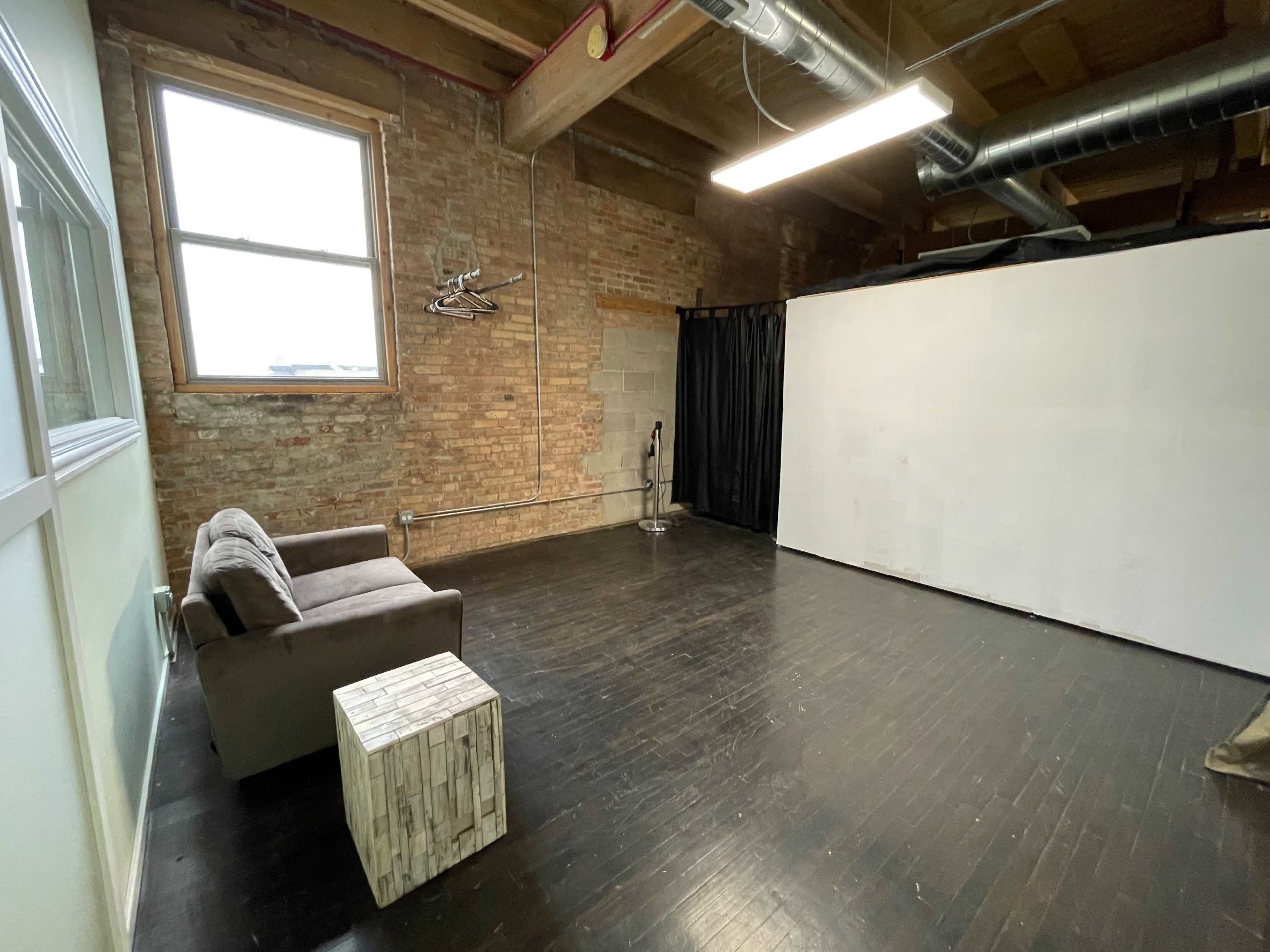 The image shows an empty industrial-style room with exposed brick walls, a wooden floor, a brown couch, a small wooden table, and a white backdrop.