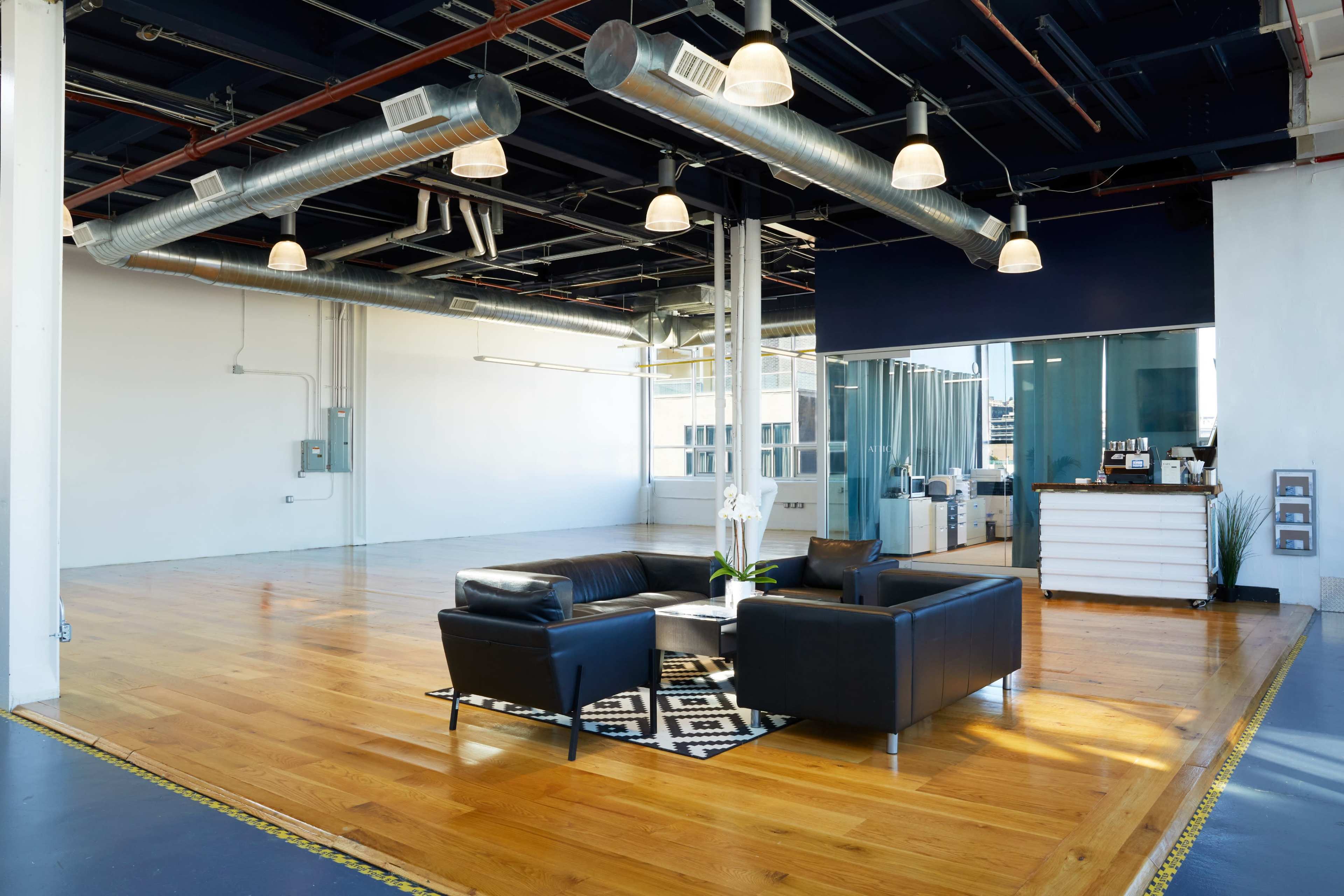 The image shows a bright, open office space featuring polished wooden floors, a black leather sofa set on a geometric rug, and modern lighting fixtures.