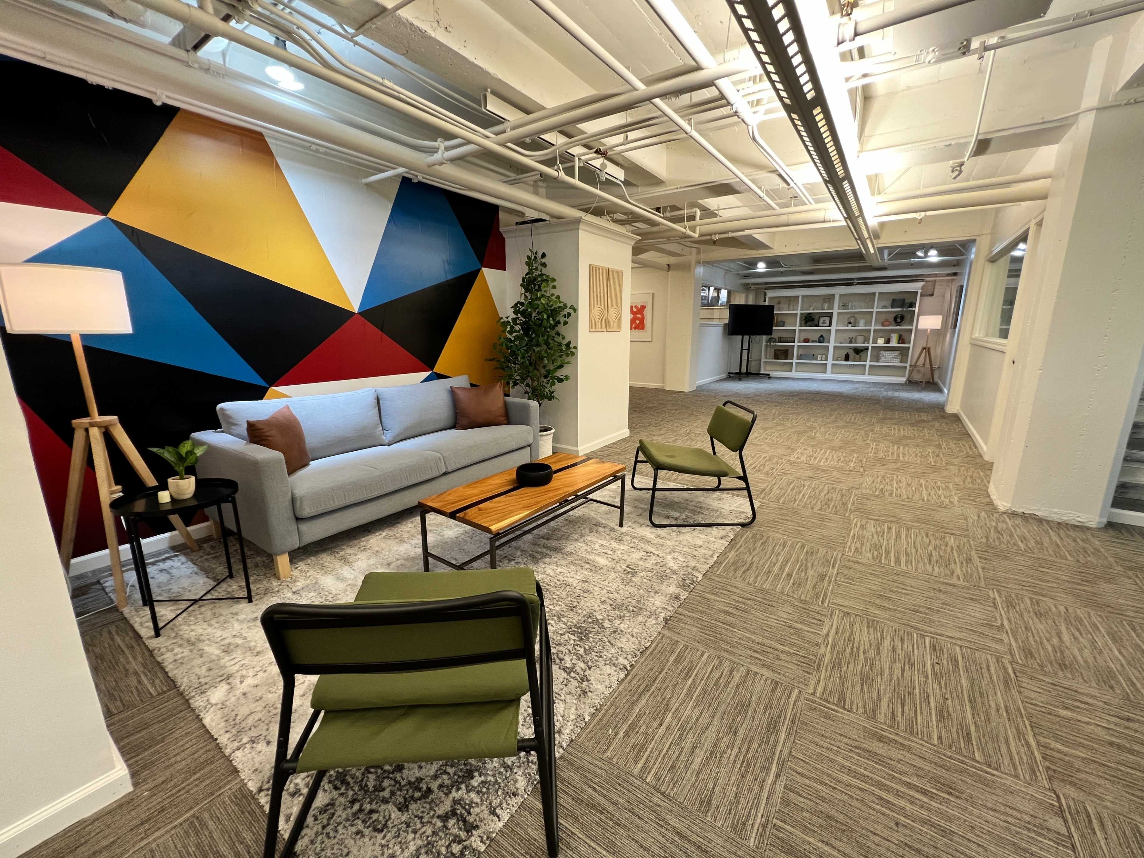 A modern lounge area with a light gray sofa, a wooden coffee table, two green chairs, and a colorful geometric wall mural.