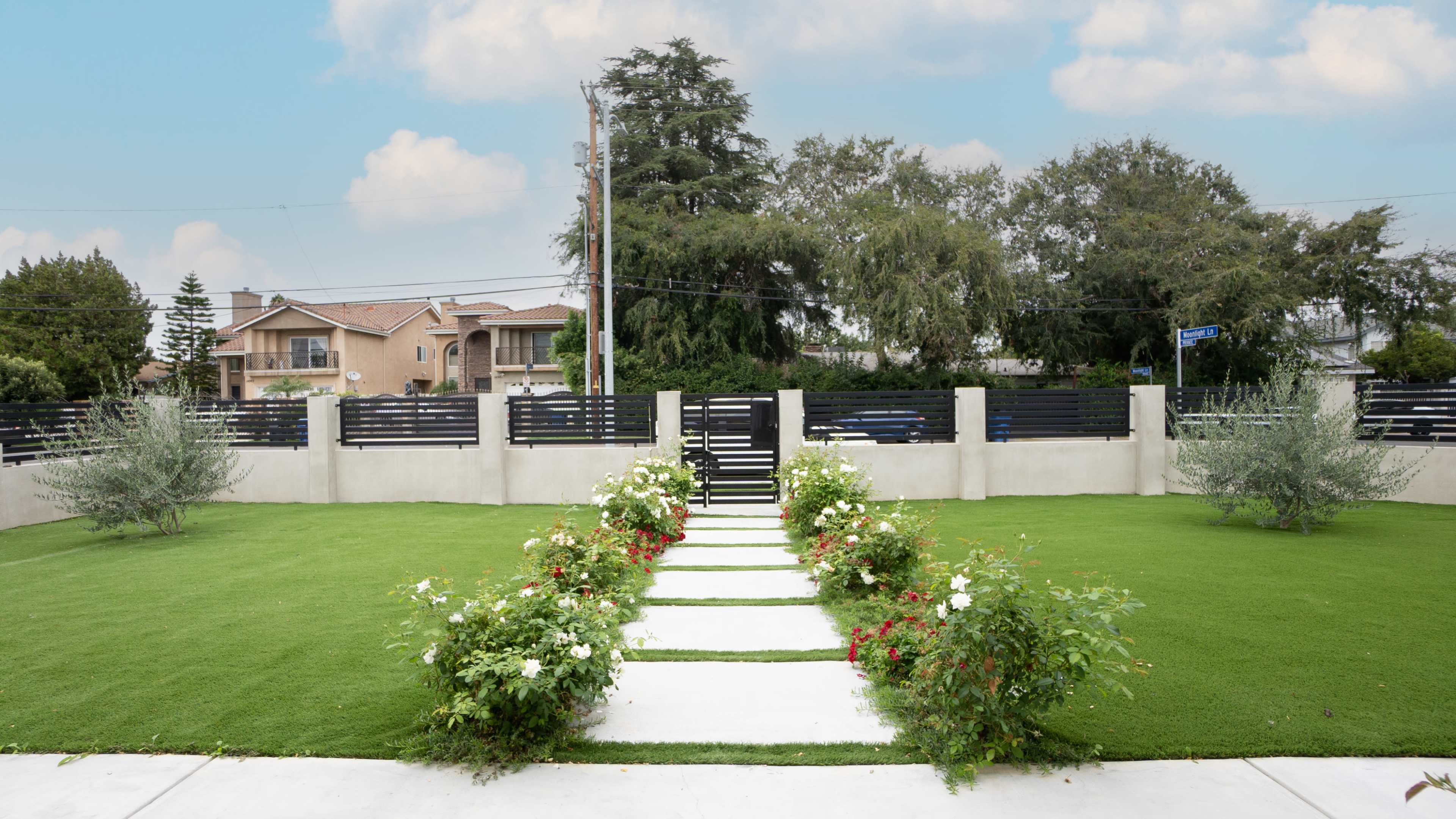 A neatly landscaped garden features a stone pathway lined with flowering bushes leading to a modern gate, with residential buildings in the background.