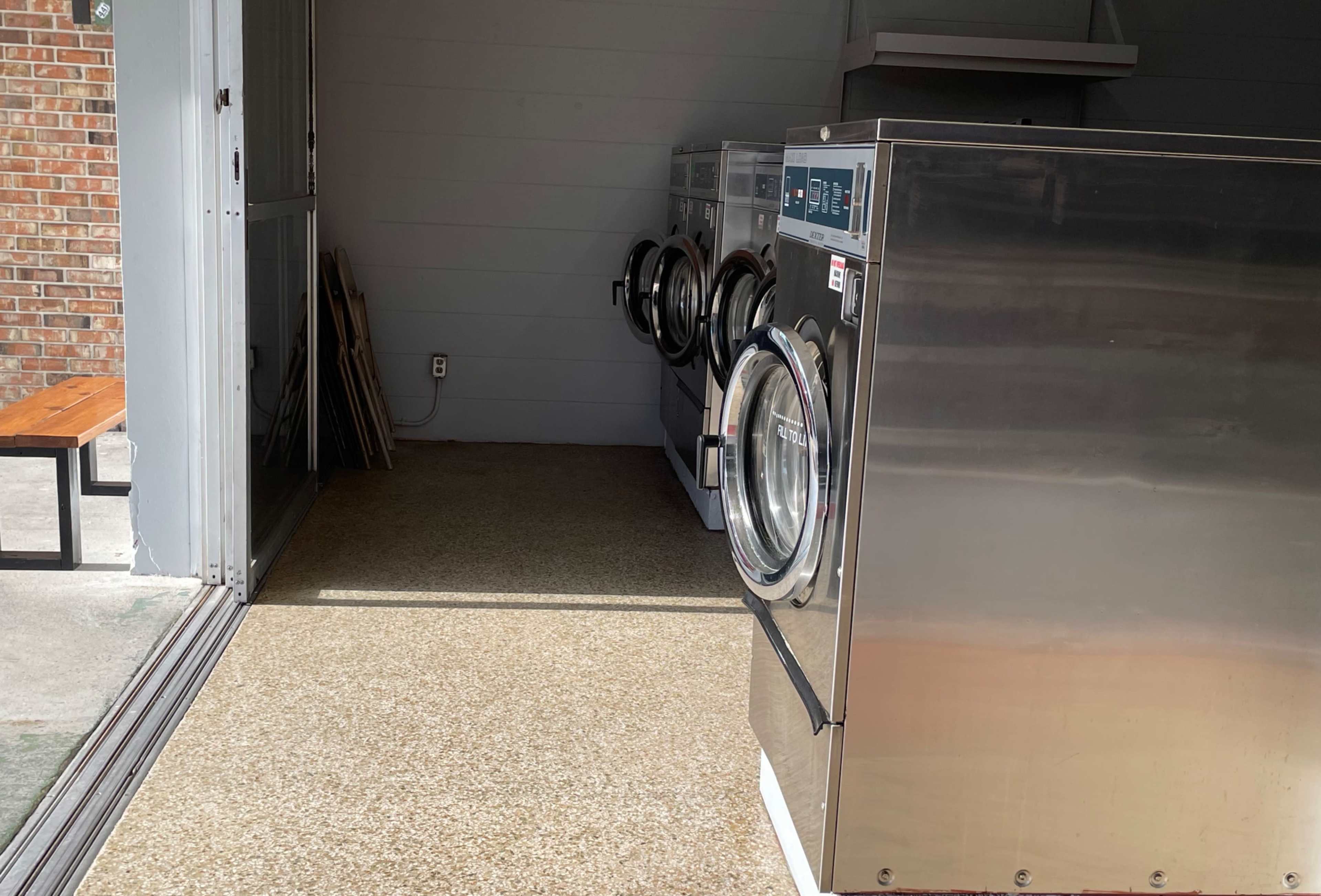 The image shows a row of commercial washing machines placed in an indoor laundry area with a concrete floor and a wooden bench in the background.
