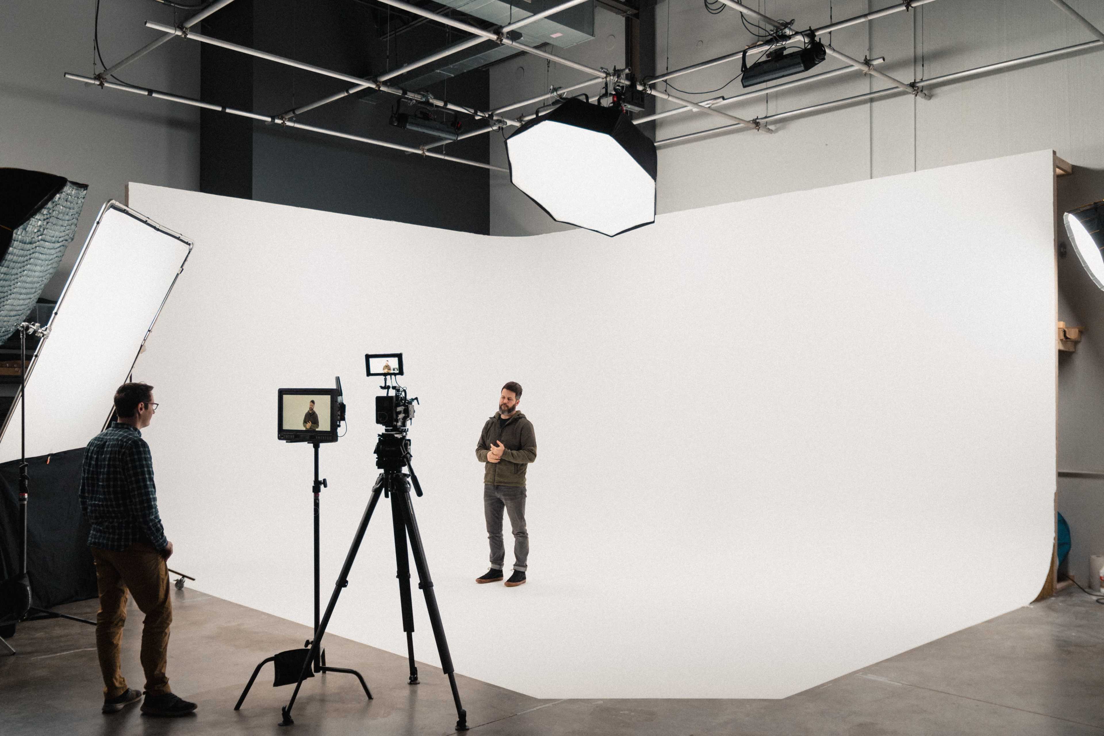 A man stands on a white backdrop speaking into a camera, while another person monitors the setup with a tablet.