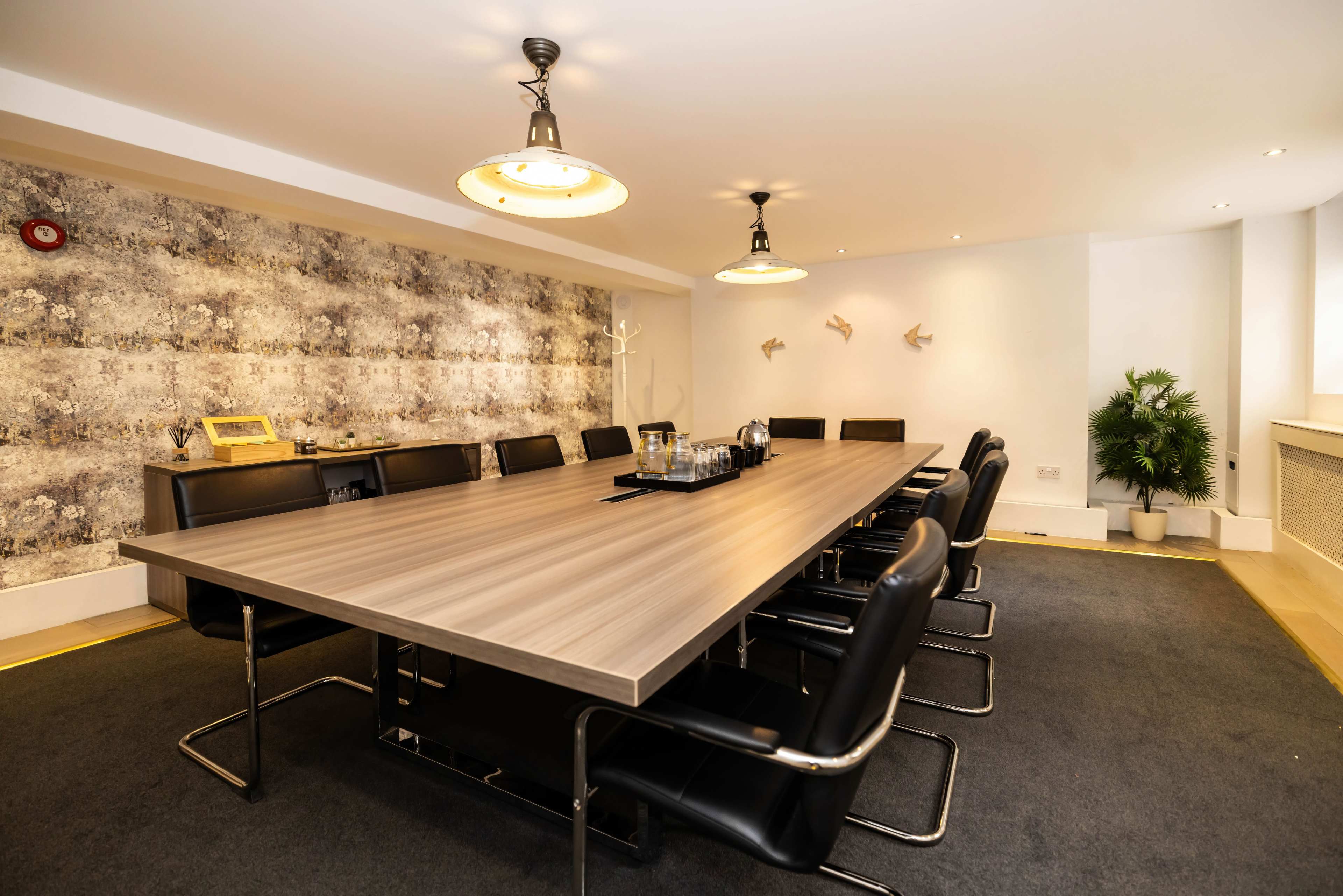 A long wooden conference table with black chairs is set in a well-lit meeting room featuring gray textured wallpaper and a potted plant.