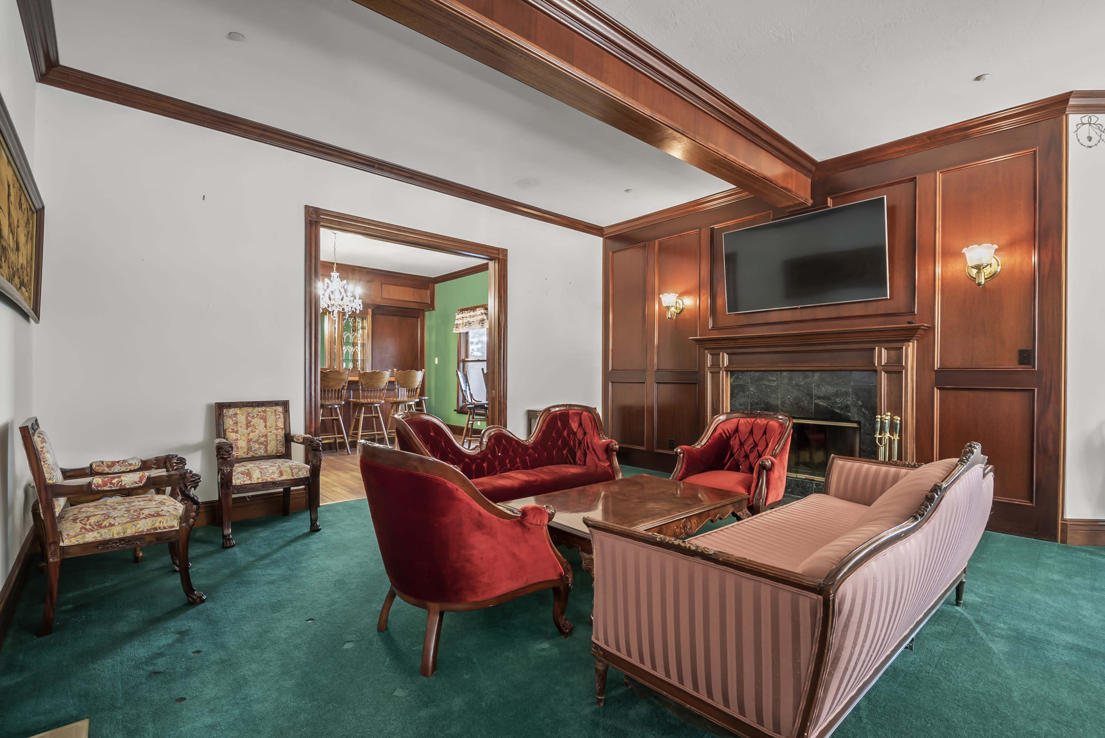 The room features a green carpet, wooden paneling, a large television mounted above a fireplace, and seating arranged with two red sofas and a pair of patterned armchairs.