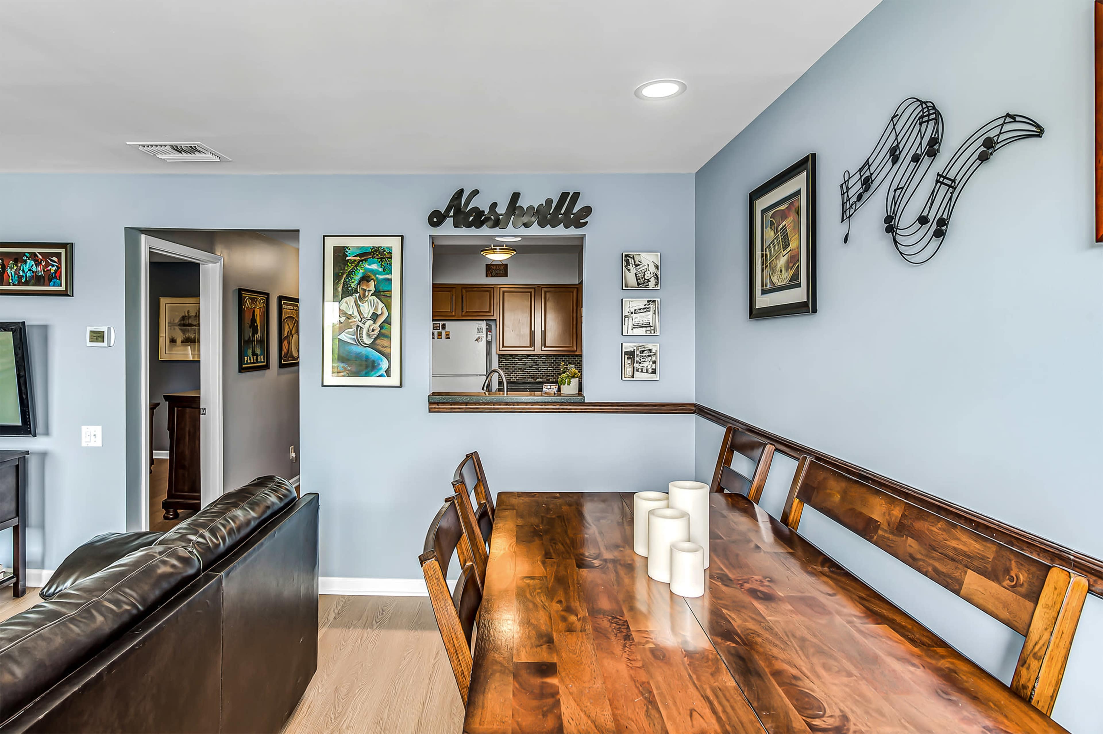 A dining area featuring a wooden table with candles, framed artwork on the walls, and a view into a kitchen area.