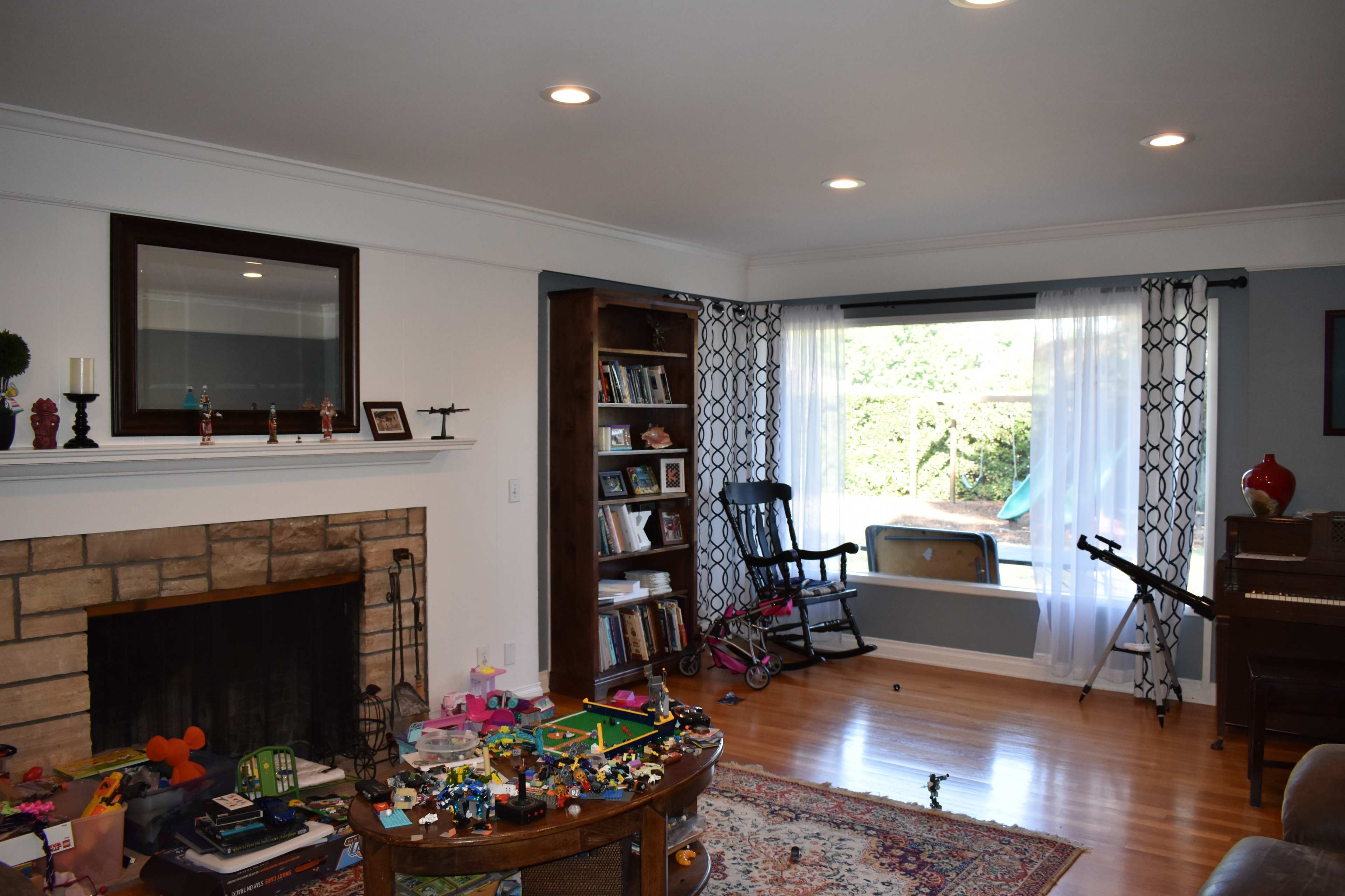 A living room features a stone fireplace, bookshelves, and scattered toys on the wooden floor, with large windows overlooking an outdoor area.