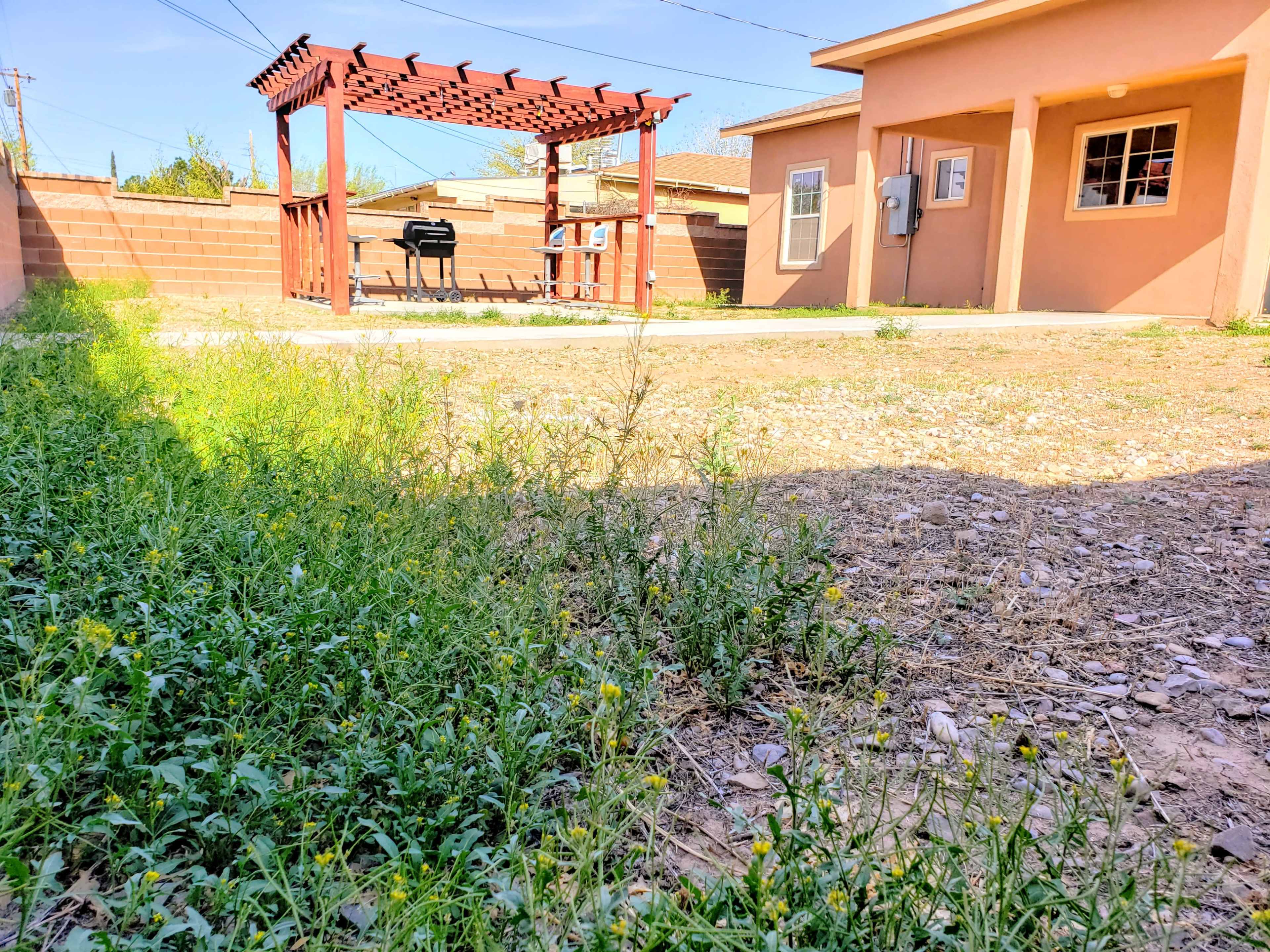 A backyard with a pergola, a grill, and a patch of grass and gravel in the foreground.