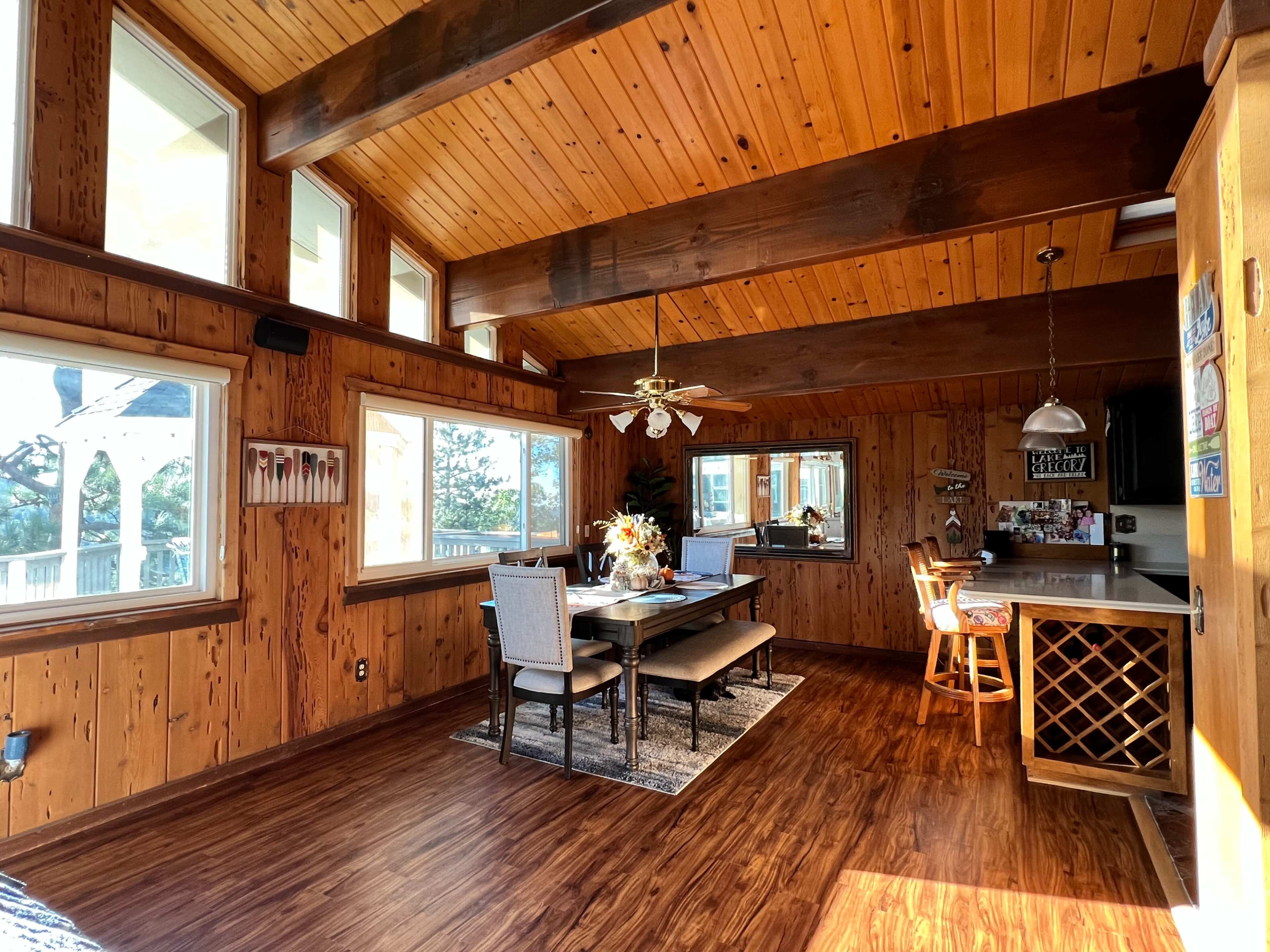 A wooden-paneled dining area with a table, chairs, a bar stool, and large windows providing natural light.