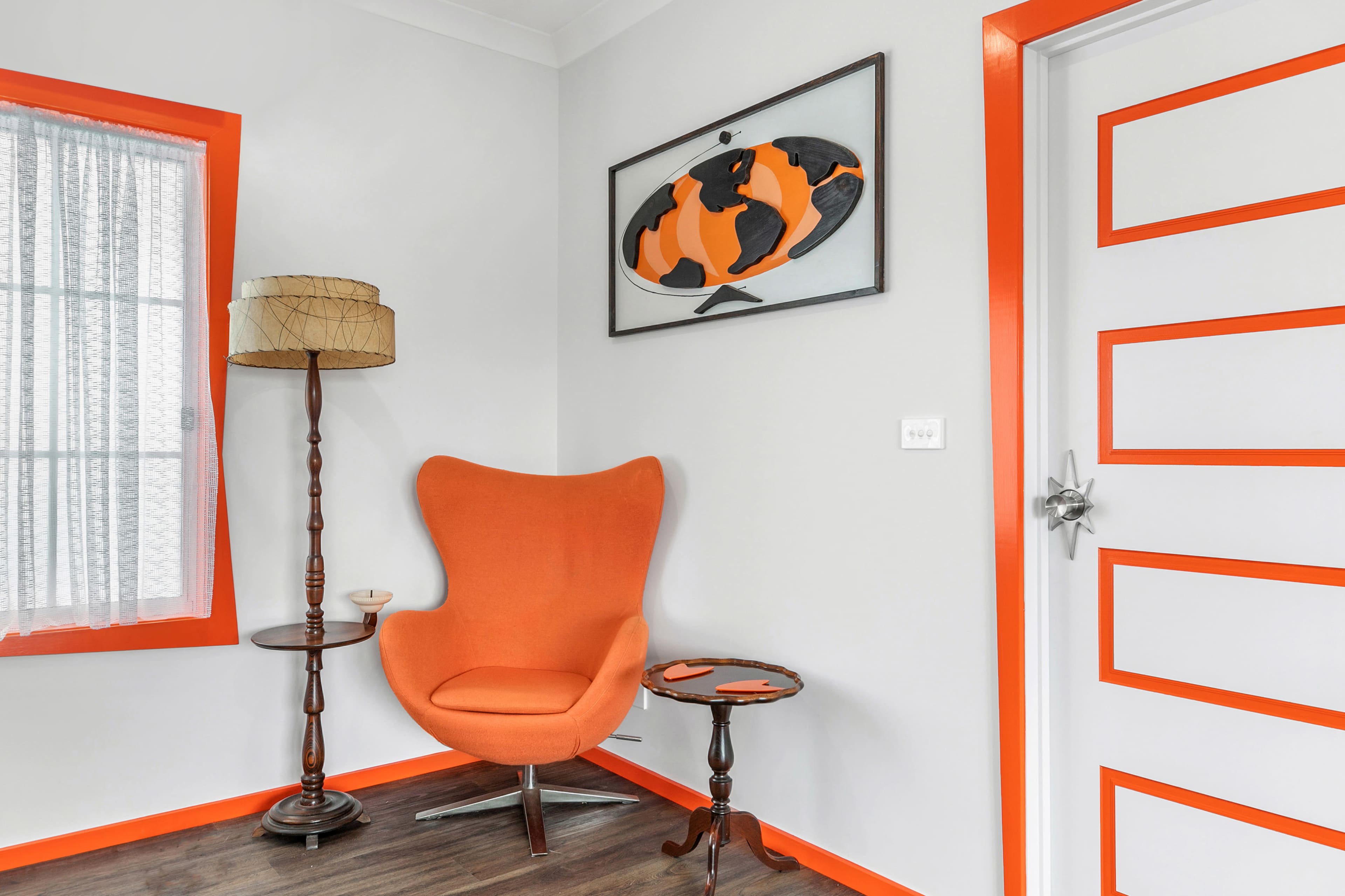 A corner of a room featuring an orange accent chair, a small side table, a floor lamp, and a door with orange trim, all set against grey walls.