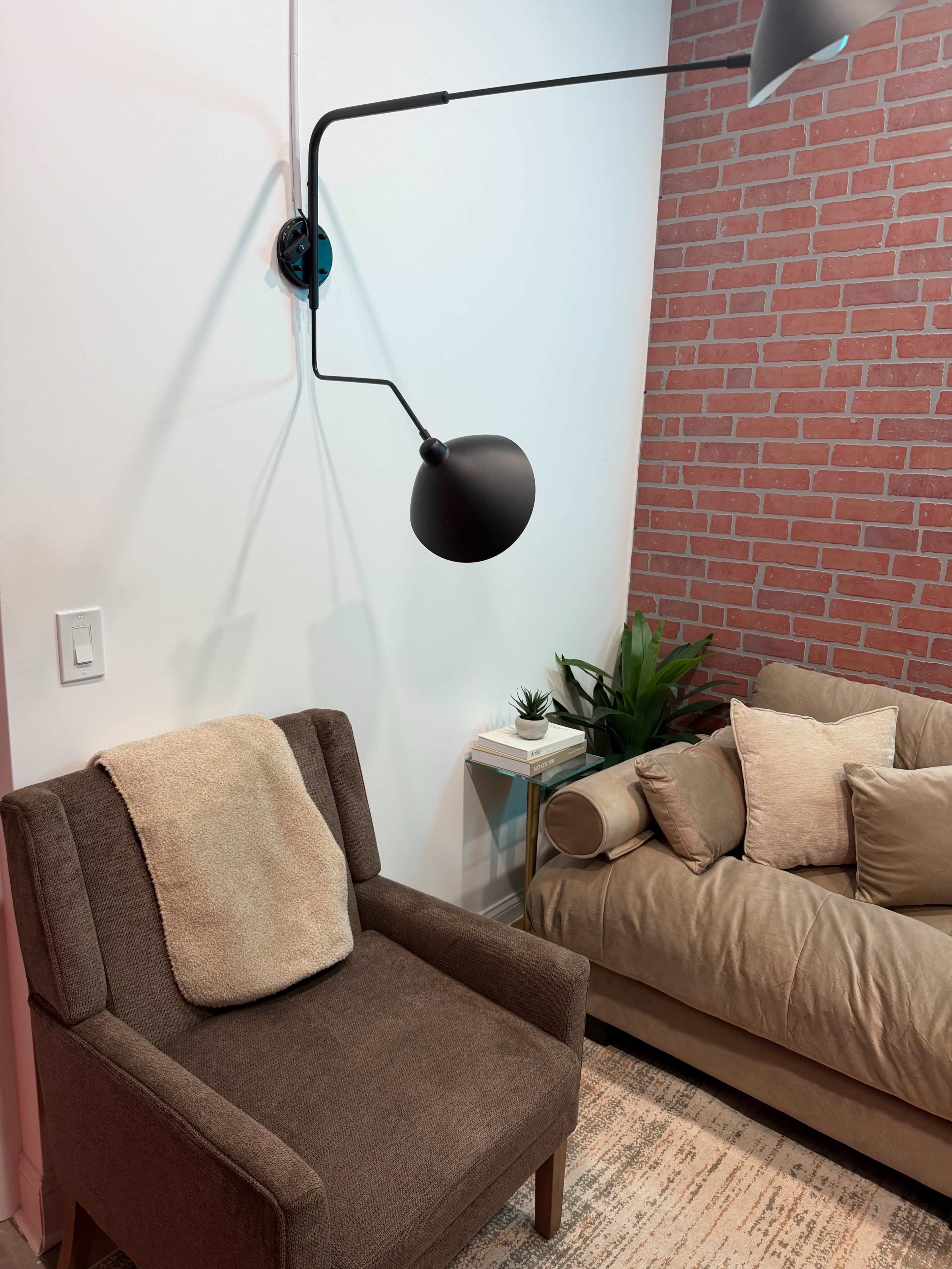 The image shows a cozy interior space featuring a brown upholstered armchair with a light-colored throw draped over it, a modern wall lamp, a small side table with books, and a sofa adorned with cushions against a brick-patterned wall.