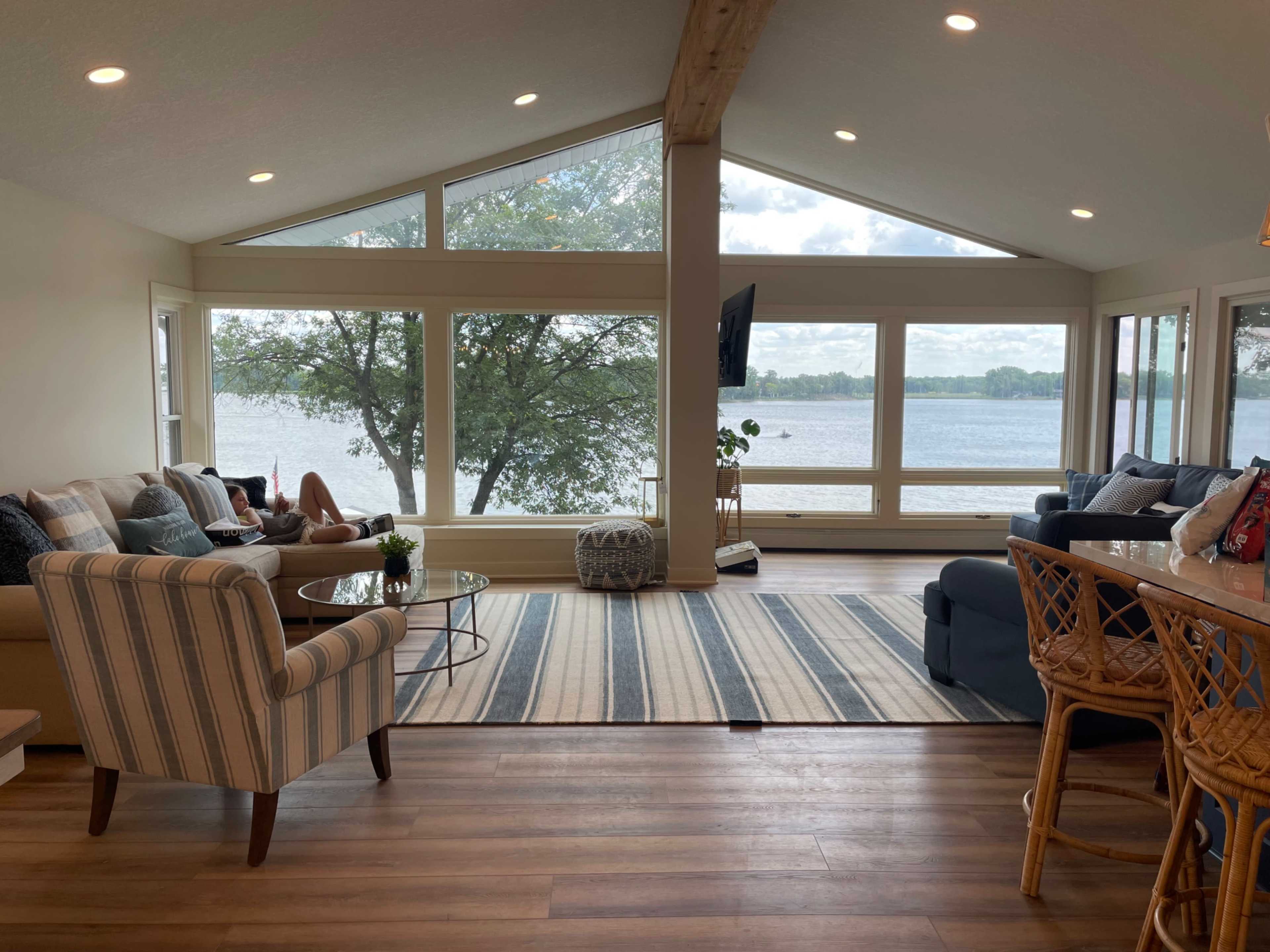 A modern living room features large windows overlooking a lake, with a comfortable seating area, a coffee table, and a striped rug.