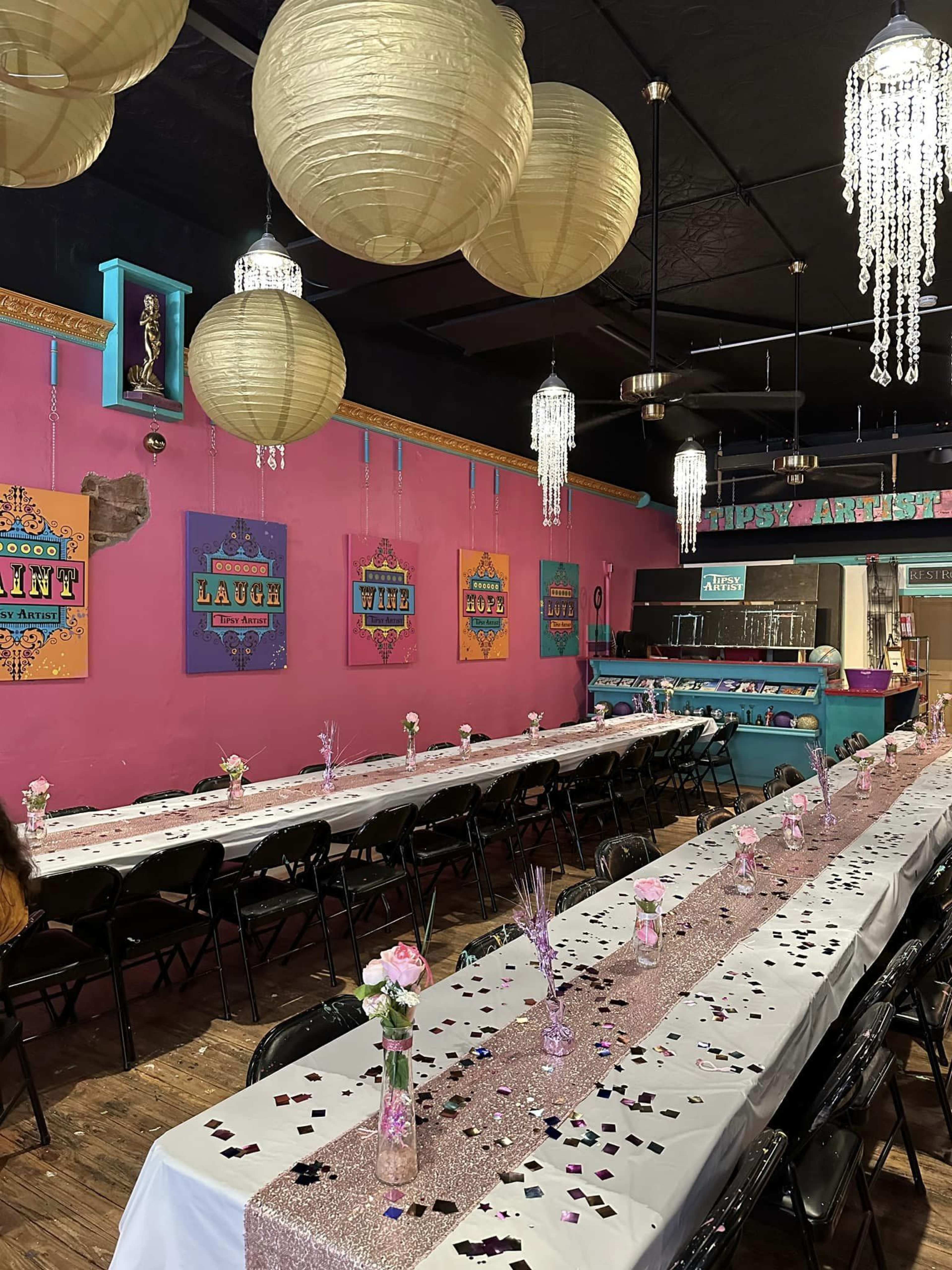 A colorful dining area is set up with long tables adorned with white tablecloths, centerpieces of flowers, and scattered confetti, surrounded by decorative lanterns and vibrant wall art.