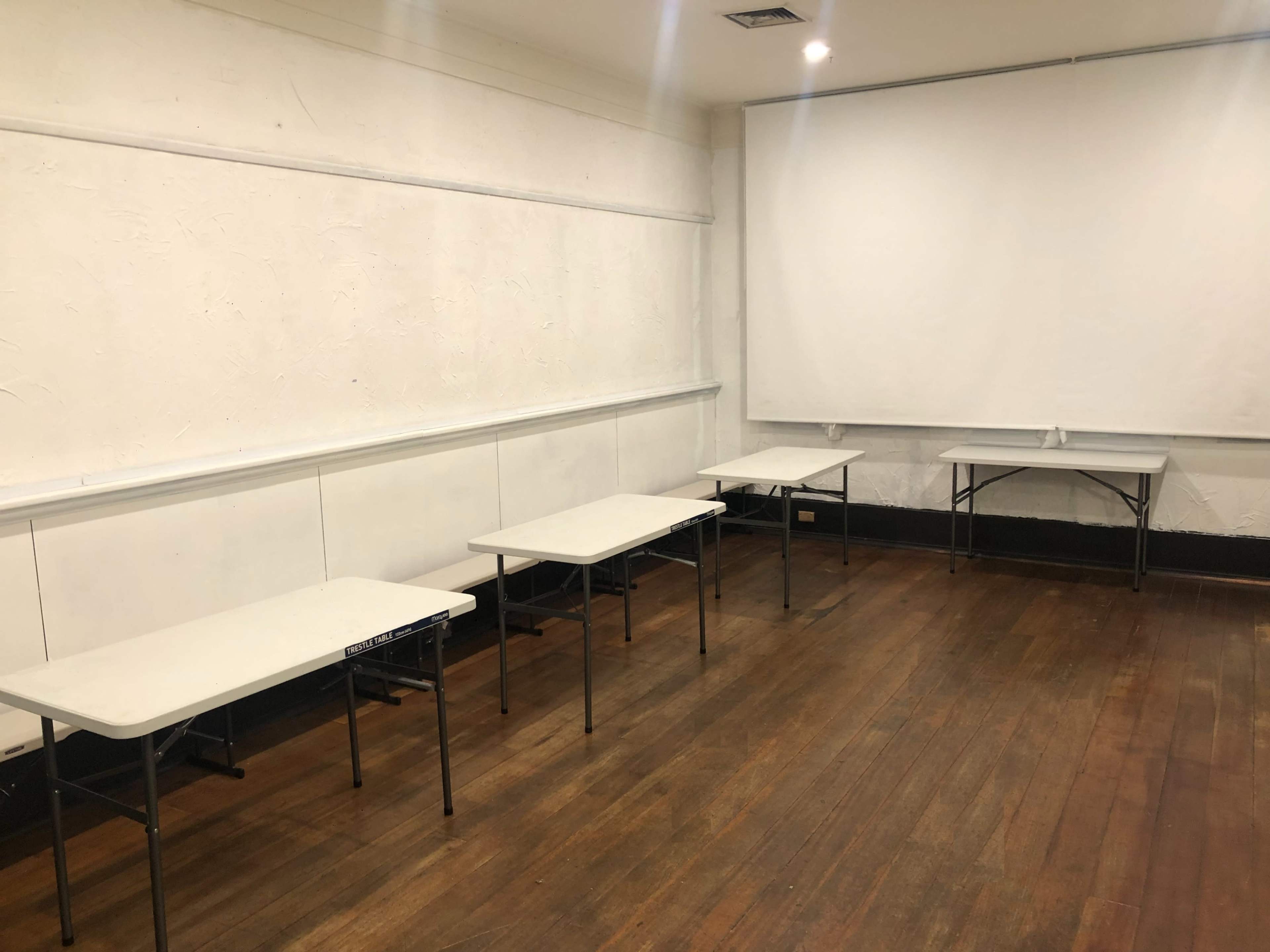 The image shows a room with wooden floors and white walls, featuring several folding tables arranged in a sparse layout.