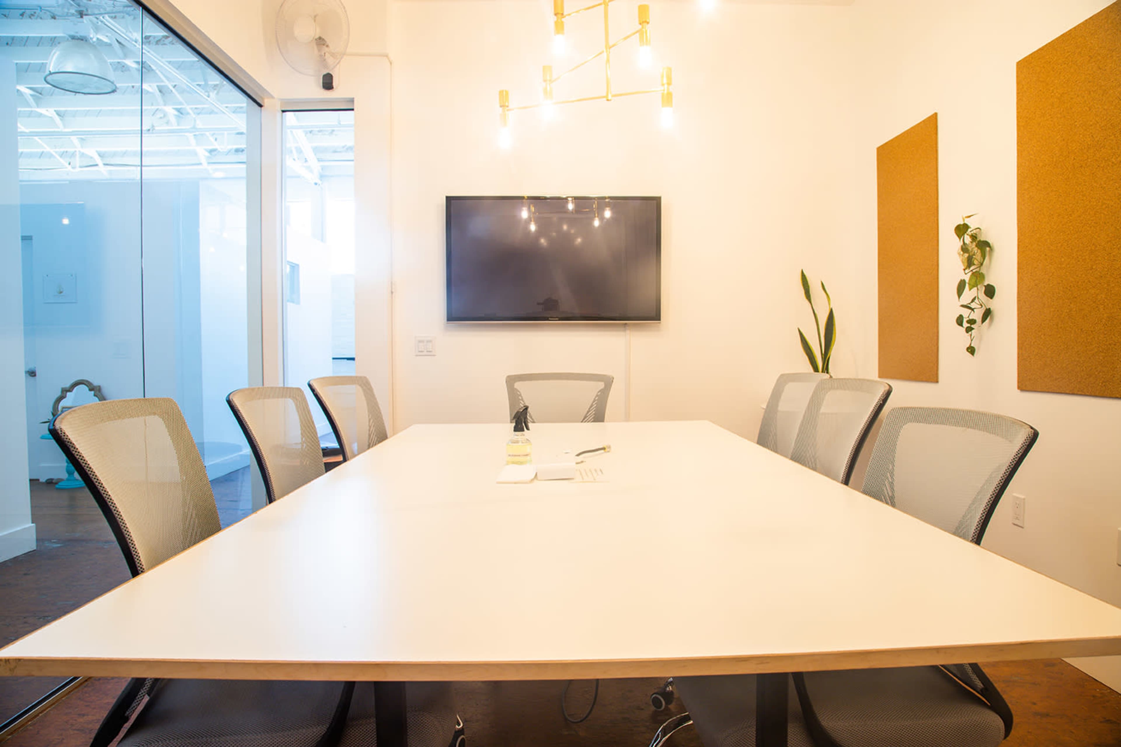 A conference room features a long white table surrounded by mesh chairs, with a TV mounted on the wall and plants in the corners.