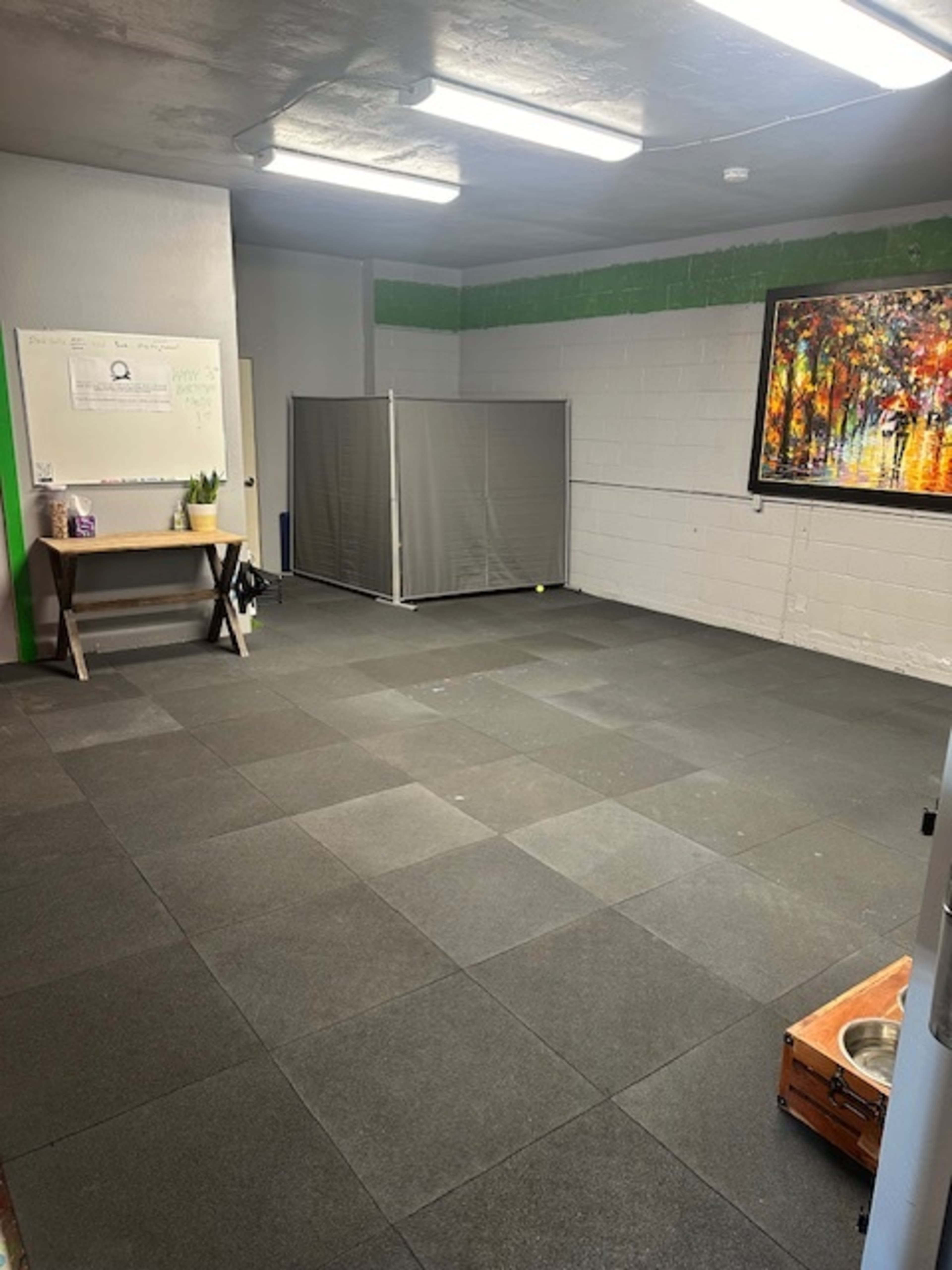 An empty room with gray tile flooring, featuring a folding partition, a table with a plant, and a colorful painting on the wall.