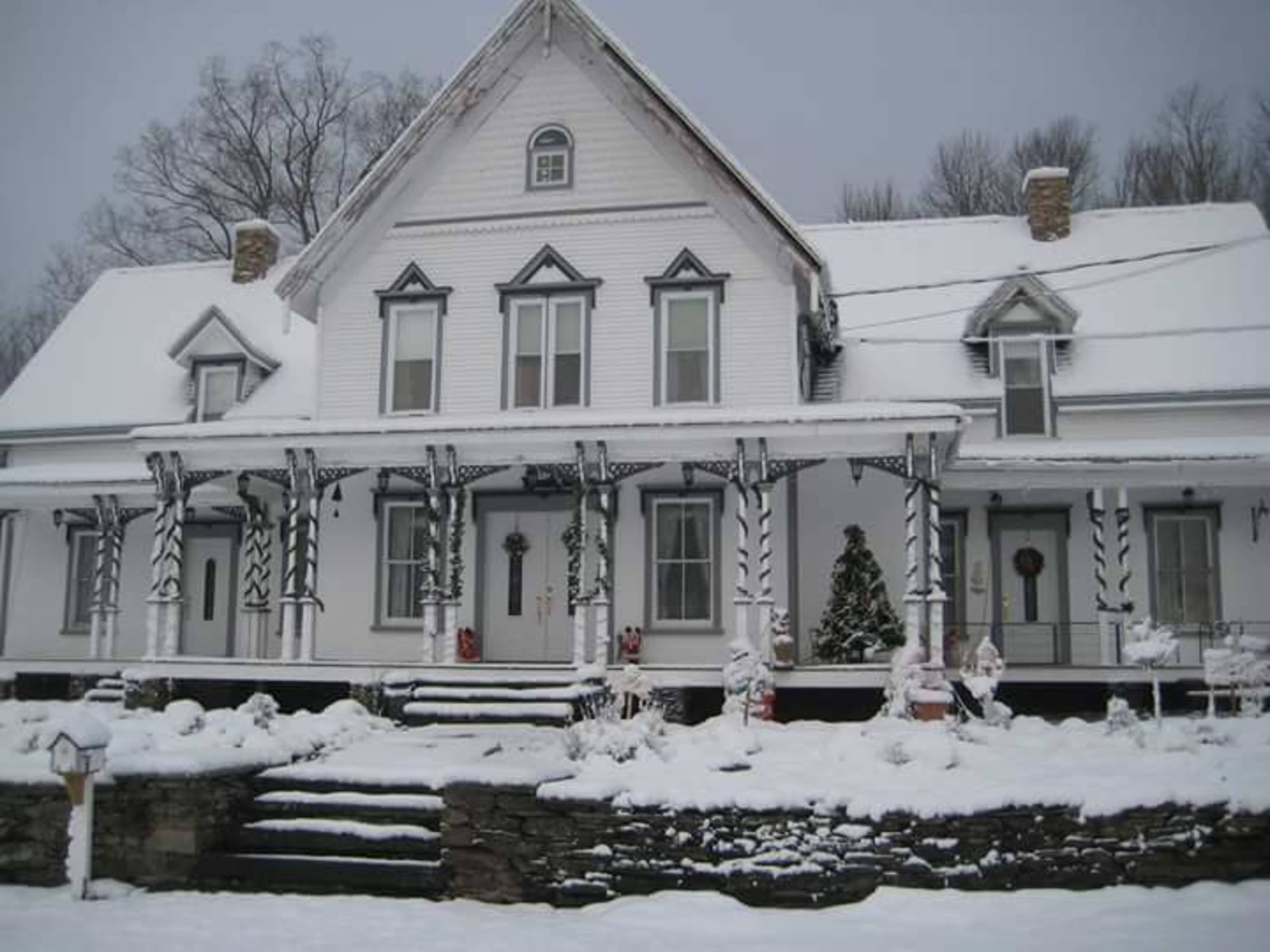 A large white house with a steep roof and a porch is surrounded by snow, featuring holiday decorations and a small evergreen tree.