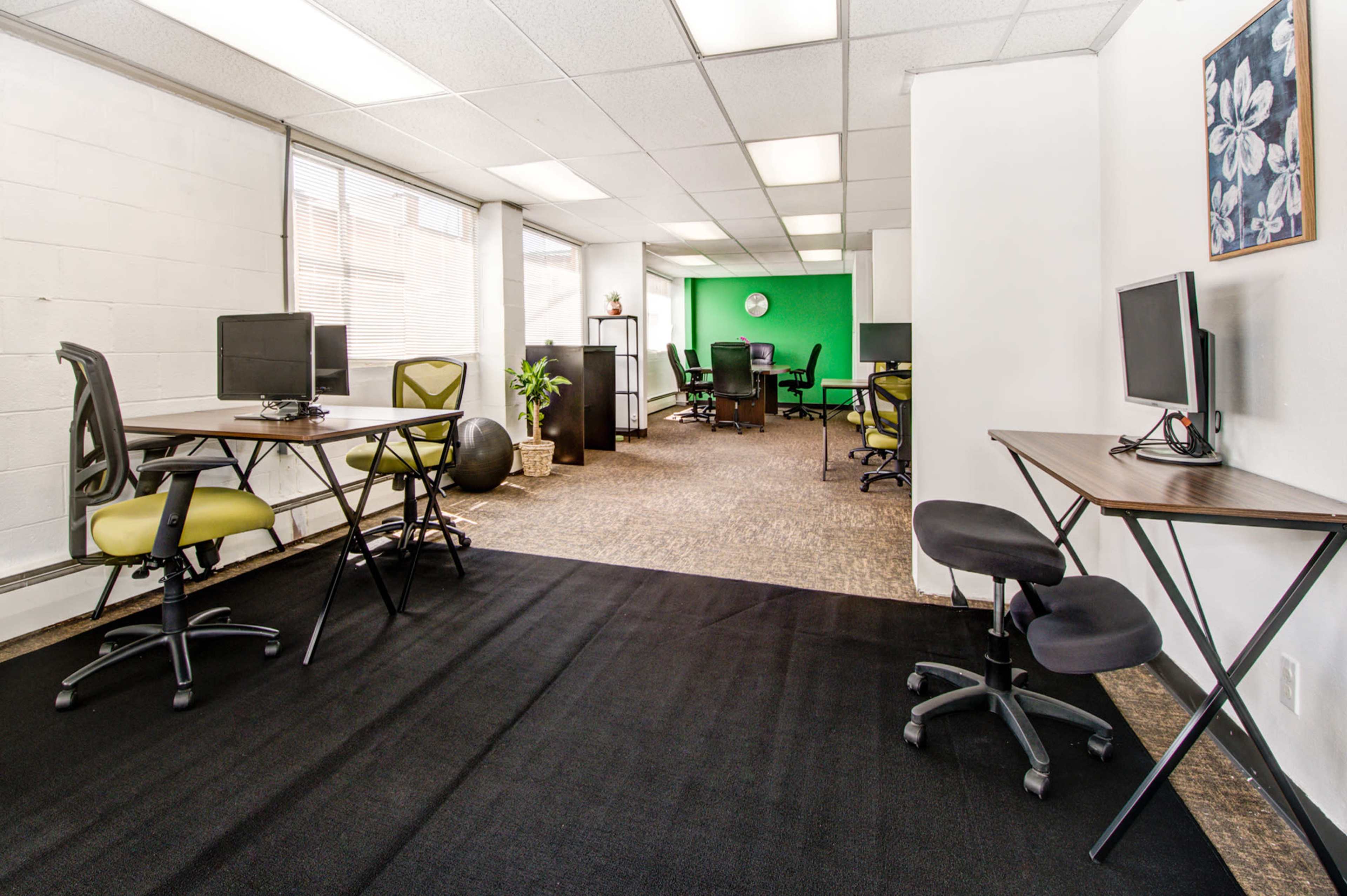 An office space with several workstations featuring chairs and computer desks, separated by a black rug and illuminated by natural light from large windows.