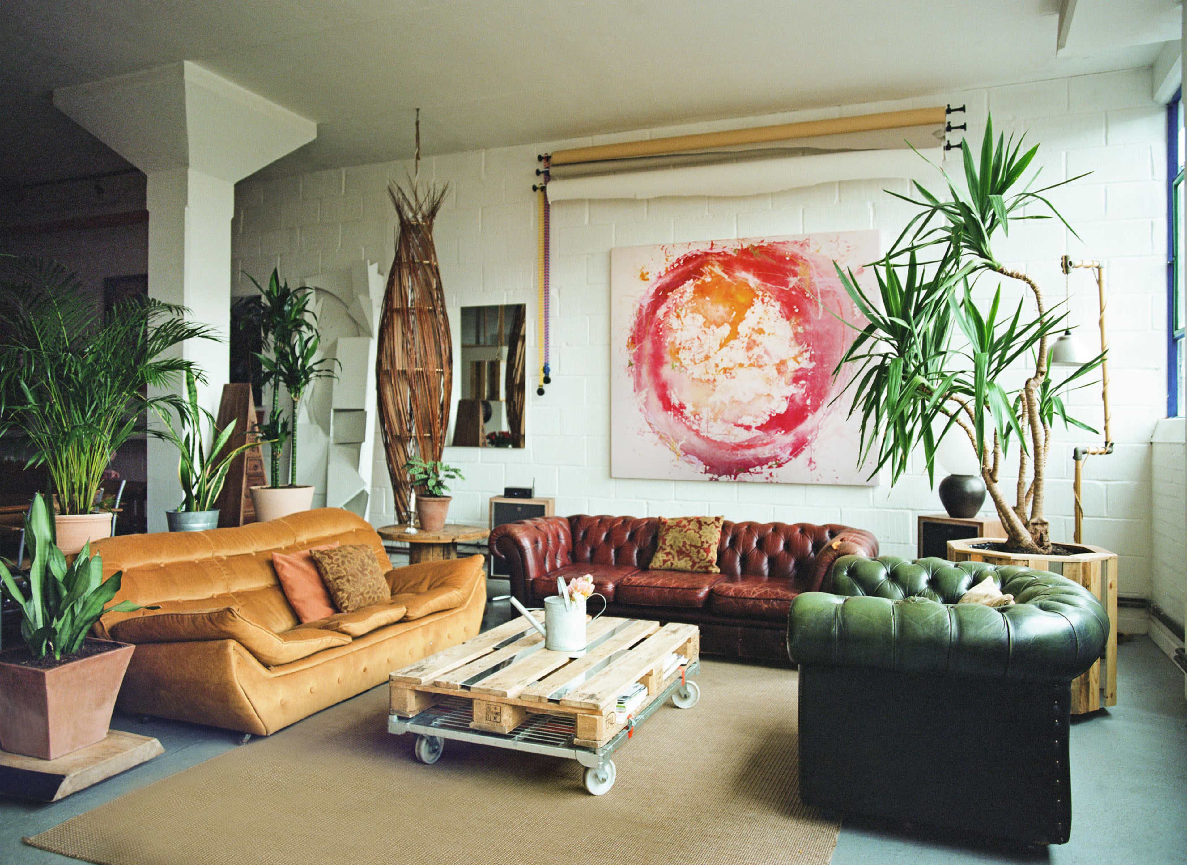 A spacious living room features a mix of brown leather sofas, a coffee table made of pallets, large indoor plants, and a colorful abstract painting on the wall.