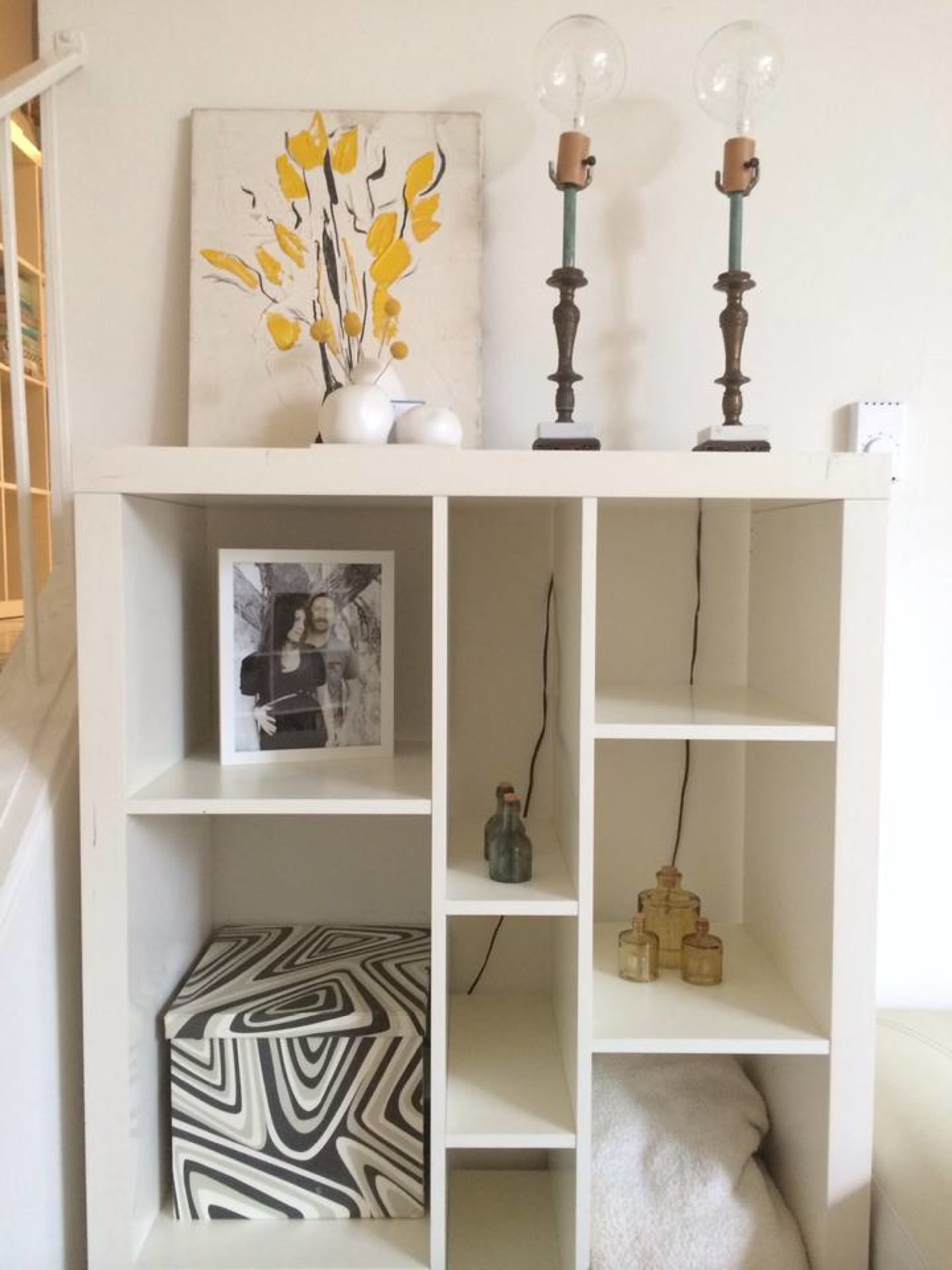 A white storage unit displays a framed photo, decorative vases, and a patterned box, with two candle holders and a painting above it.