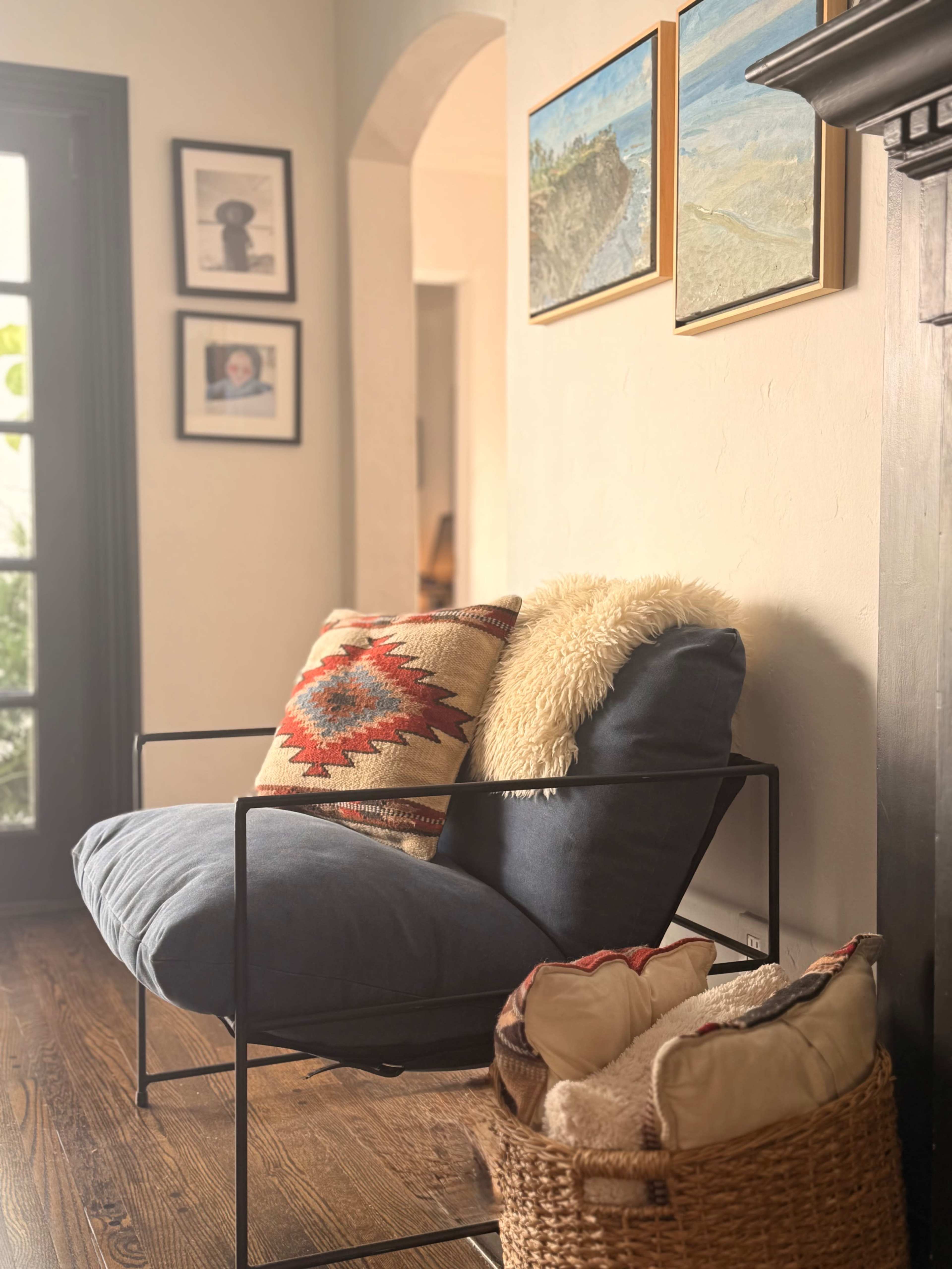 The image shows a cozy seating area featuring a blue and gray chair with a decorative pillow and a faux fur throw, alongside a basket filled with blankets and a wall adorned with framed artwork and photos.
