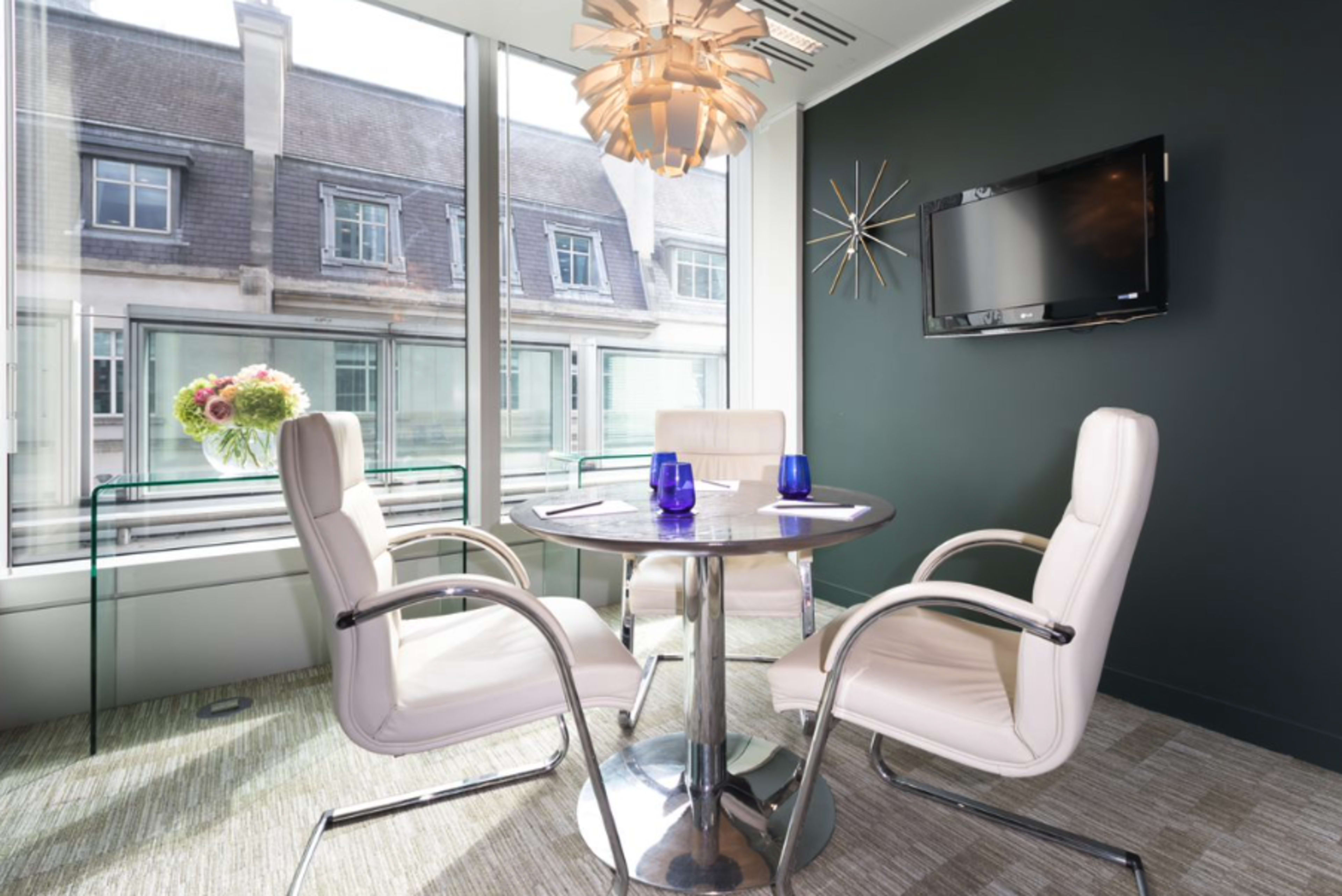 A modern meeting room features a round table with four white chairs, large windows overlooking a building exterior, and decorative elements including a pendant light and a wall-mounted TV.