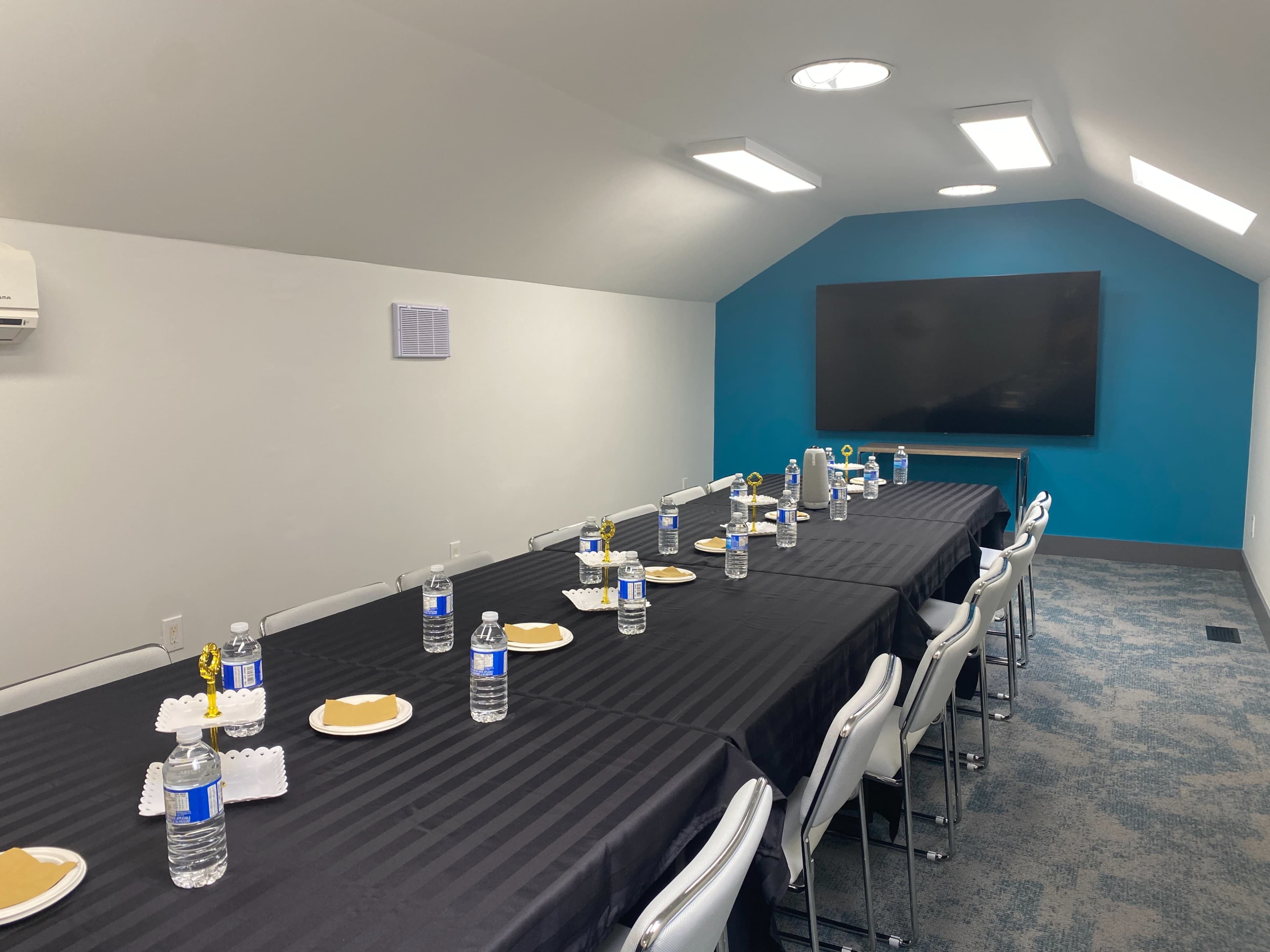 A conference room is set up with a long table covered in a black tablecloth, featuring bottles of water and plates arranged along its length, with a large screen on the wall at one end.