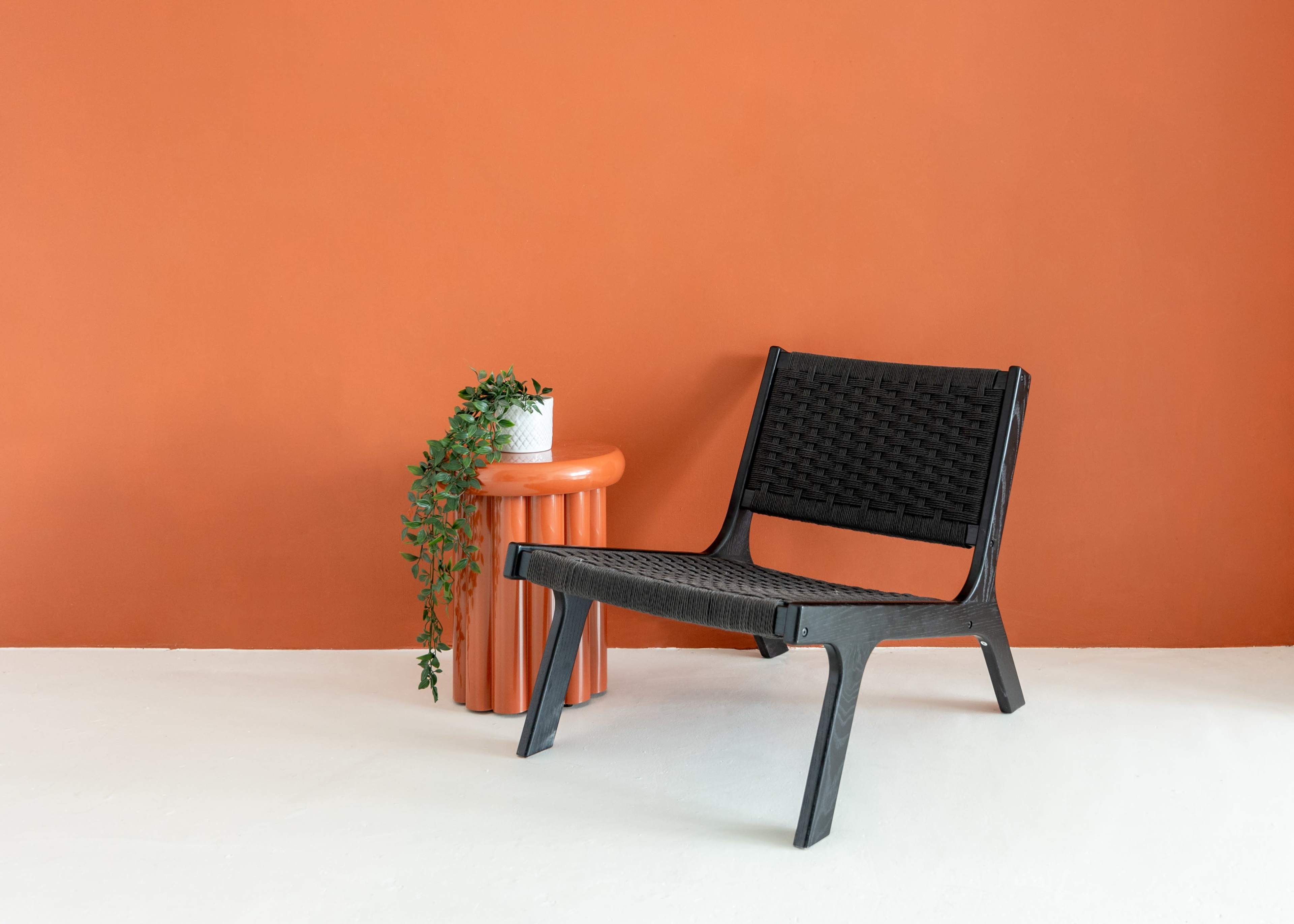 A black woven chair is positioned next to a round orange side table with a potted plant on top, against an orange wall.