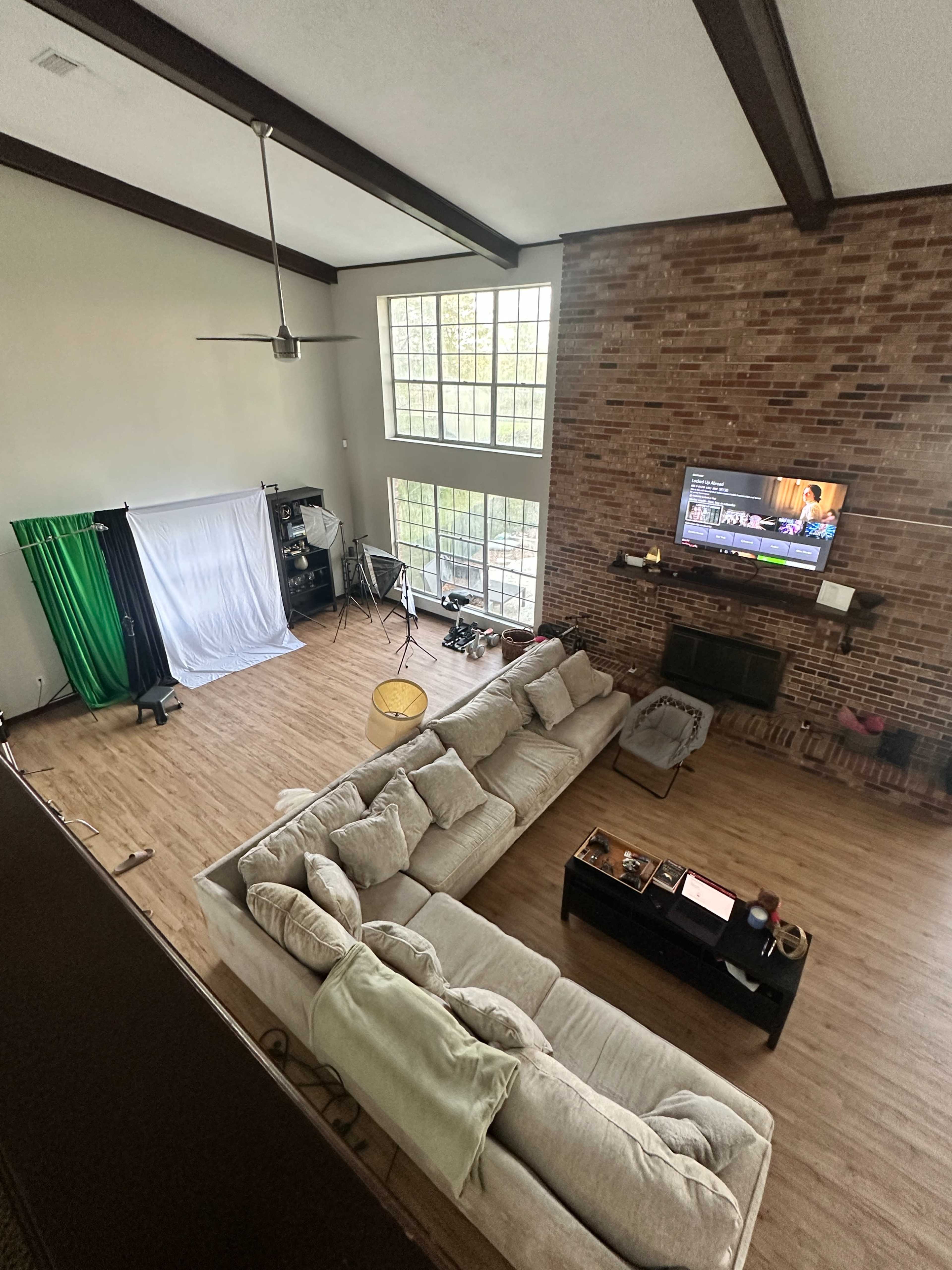 The image shows a spacious living room with a large sectional sofa, a brick wall, a television mounted above a fireplace, and a backdrop setup for photography in one corner.