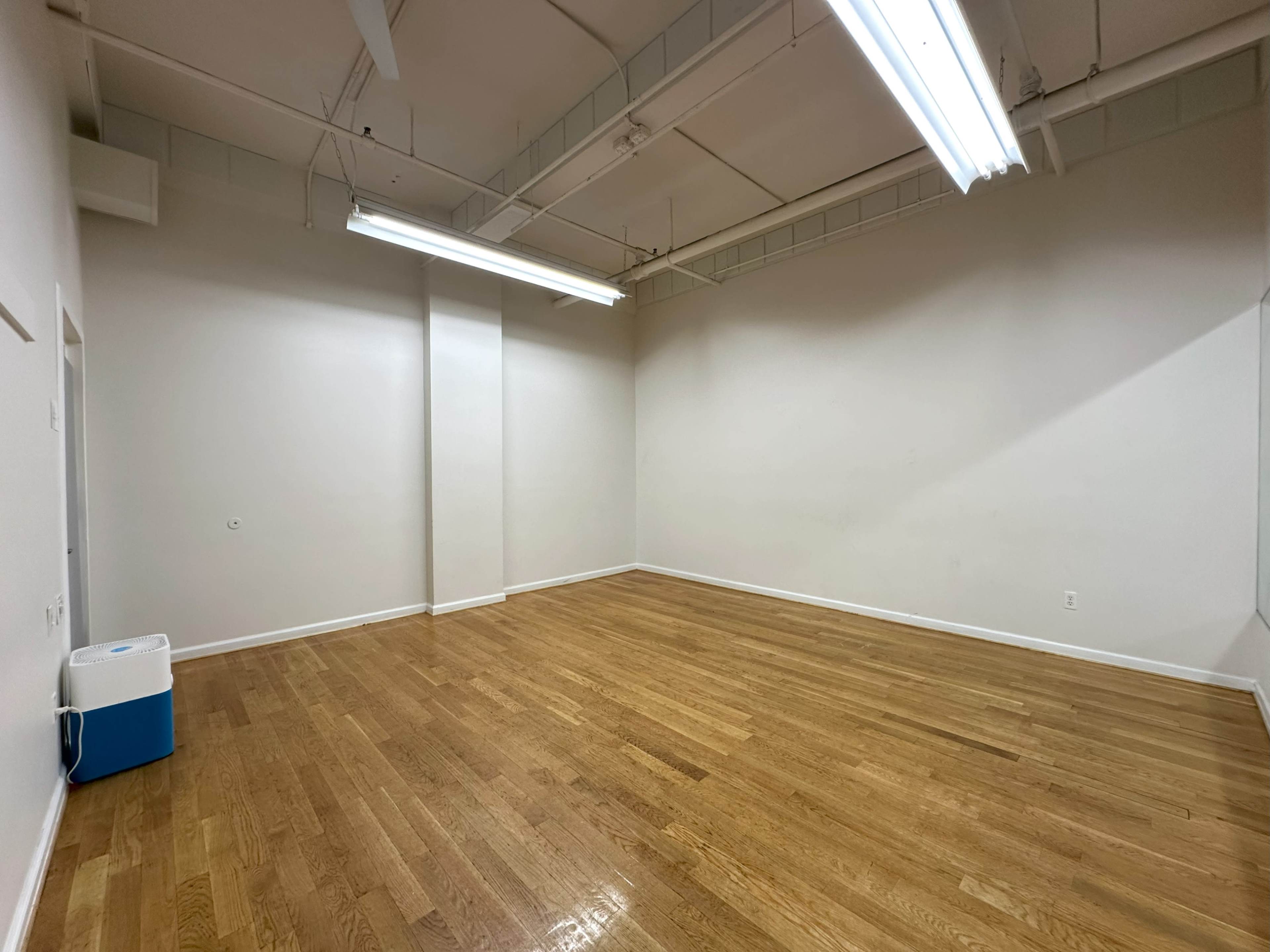 The image shows an empty room with wooden flooring, white walls, and a cooler in the corner.