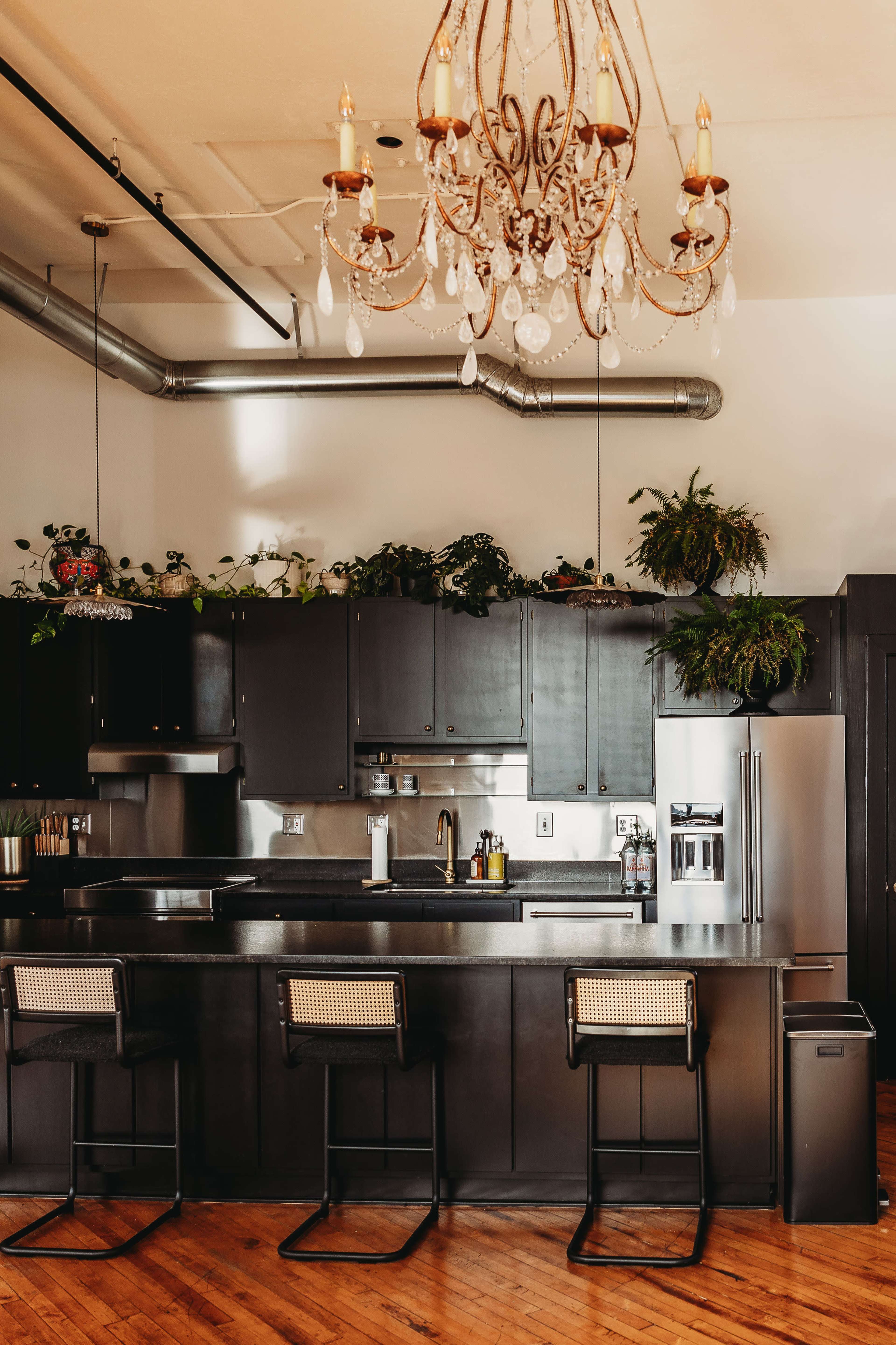 The image shows a modern kitchen with dark cabinetry, a large island with bar stools, and a chandelier overhead.