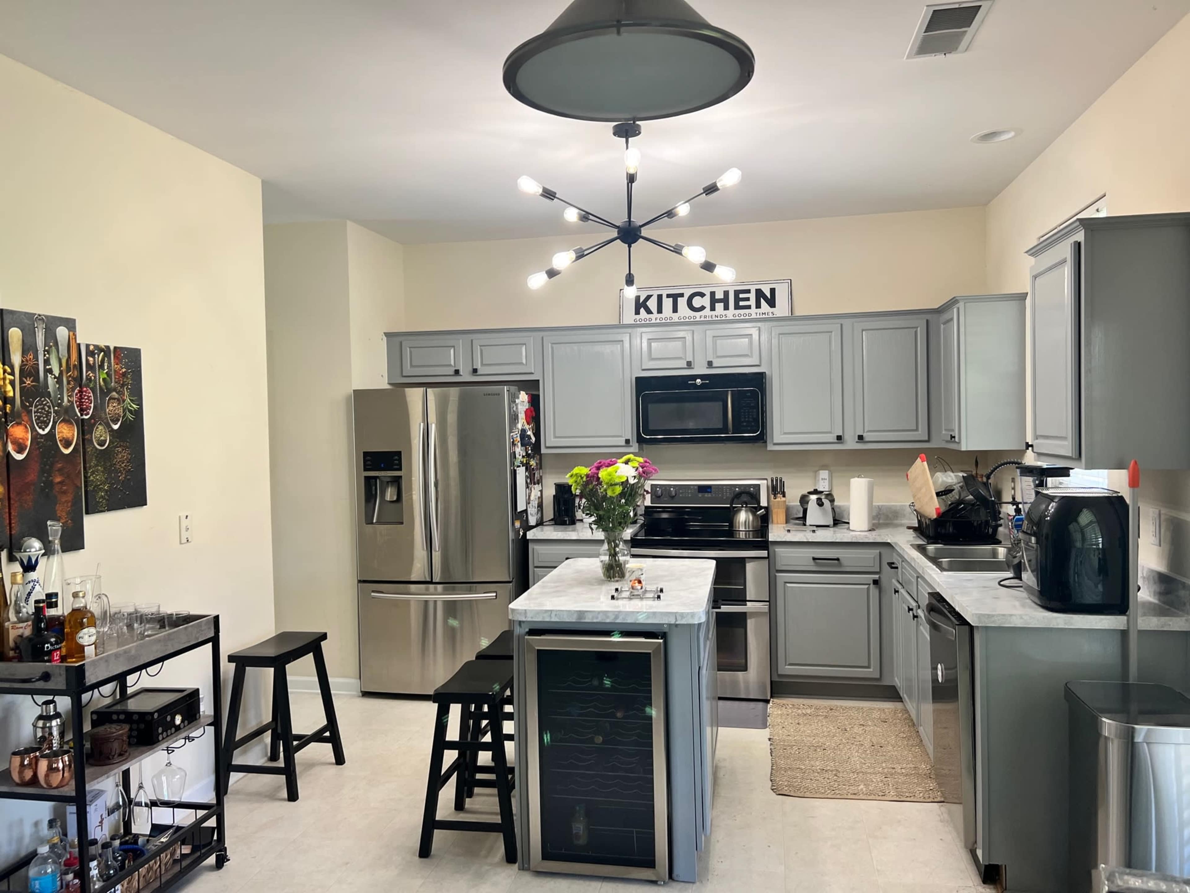 A modern kitchen features gray cabinets, stainless steel appliances, and a central island with seating, illuminated by a decorative light fixture.