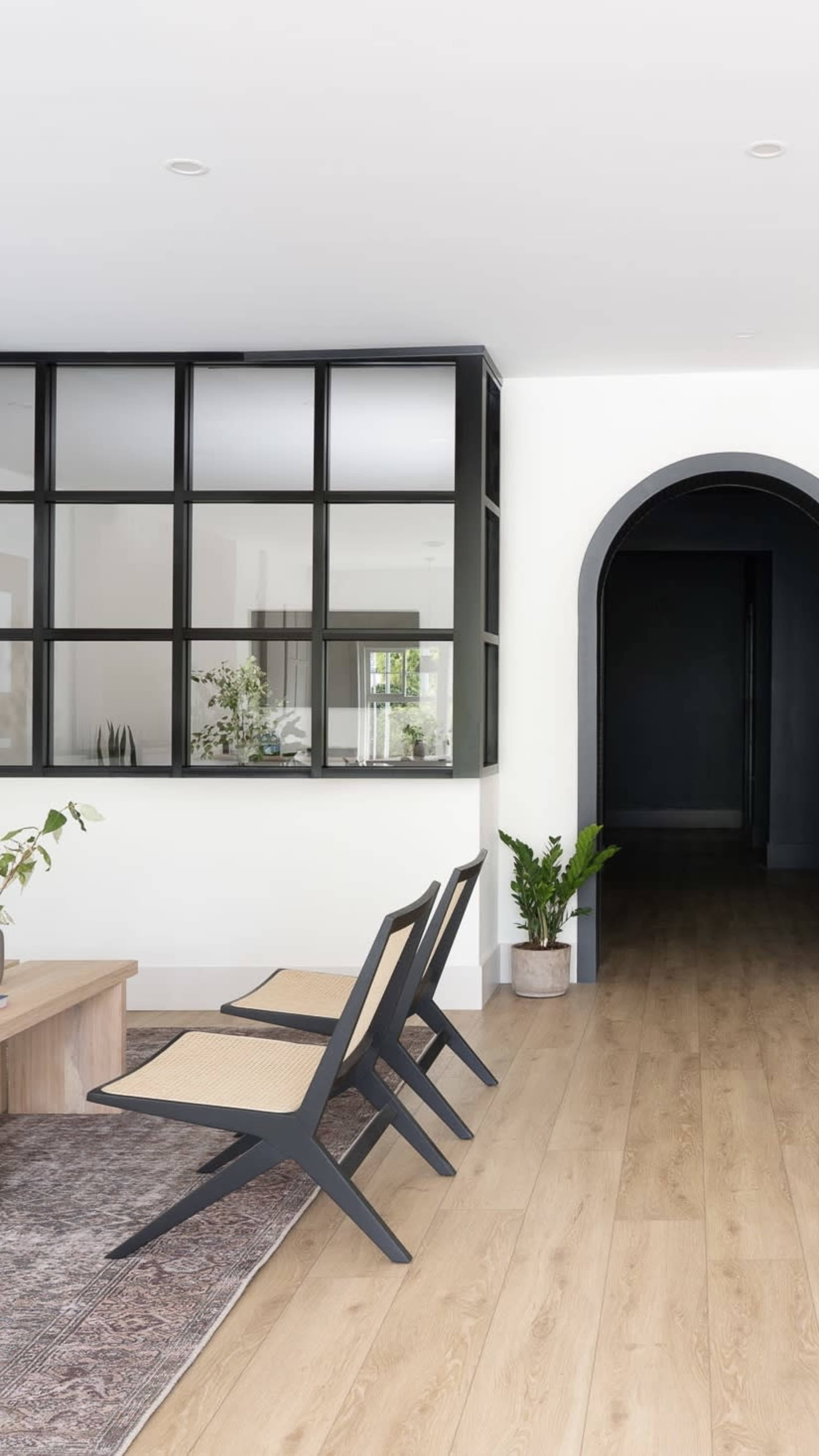 A minimalist interior features two black chairs with woven seats in front of a glass partition, leading to a dark hallway.