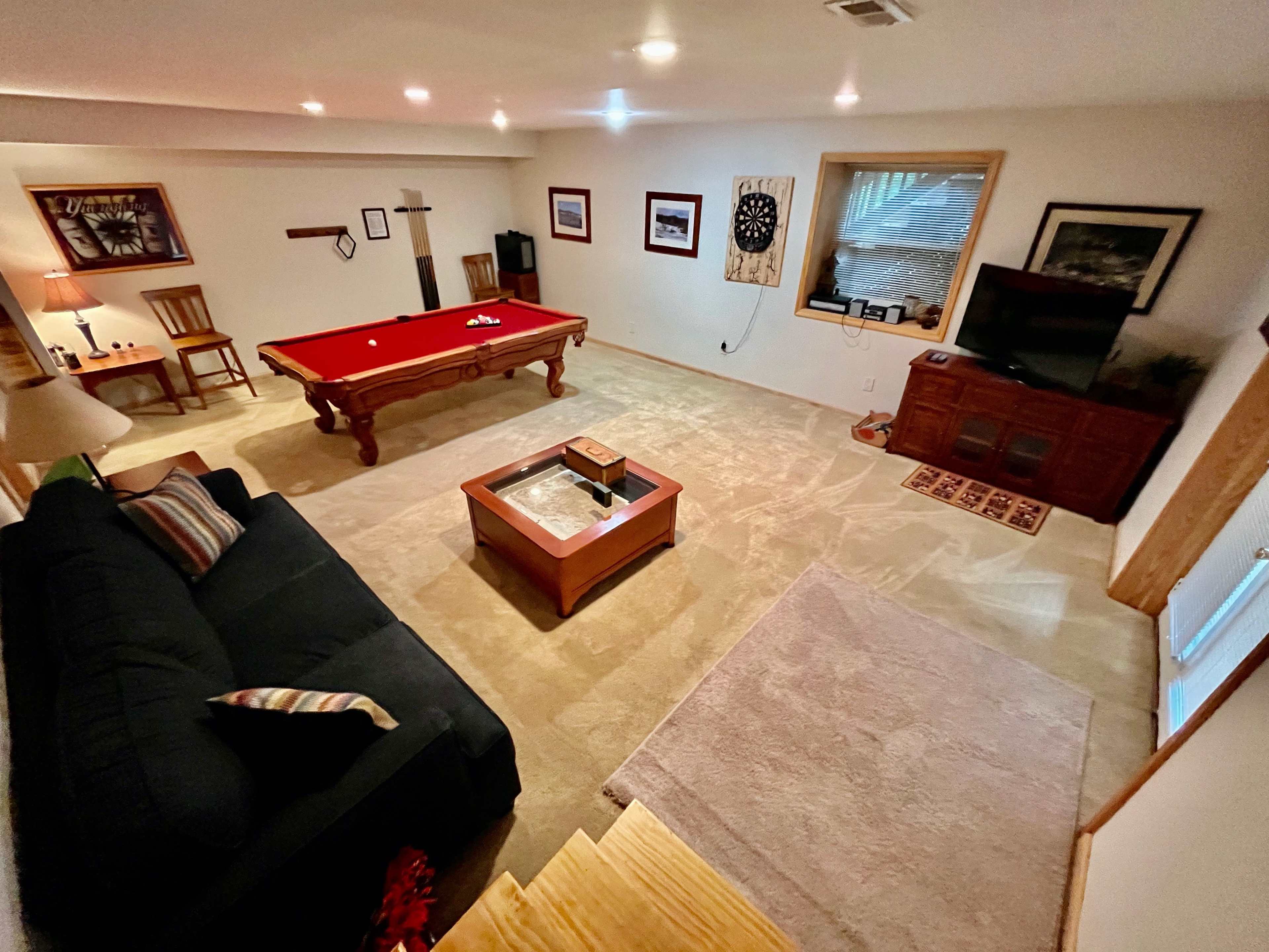 A spacious basement recreation room featuring a pool table, a dark couch, a coffee table, and a television.