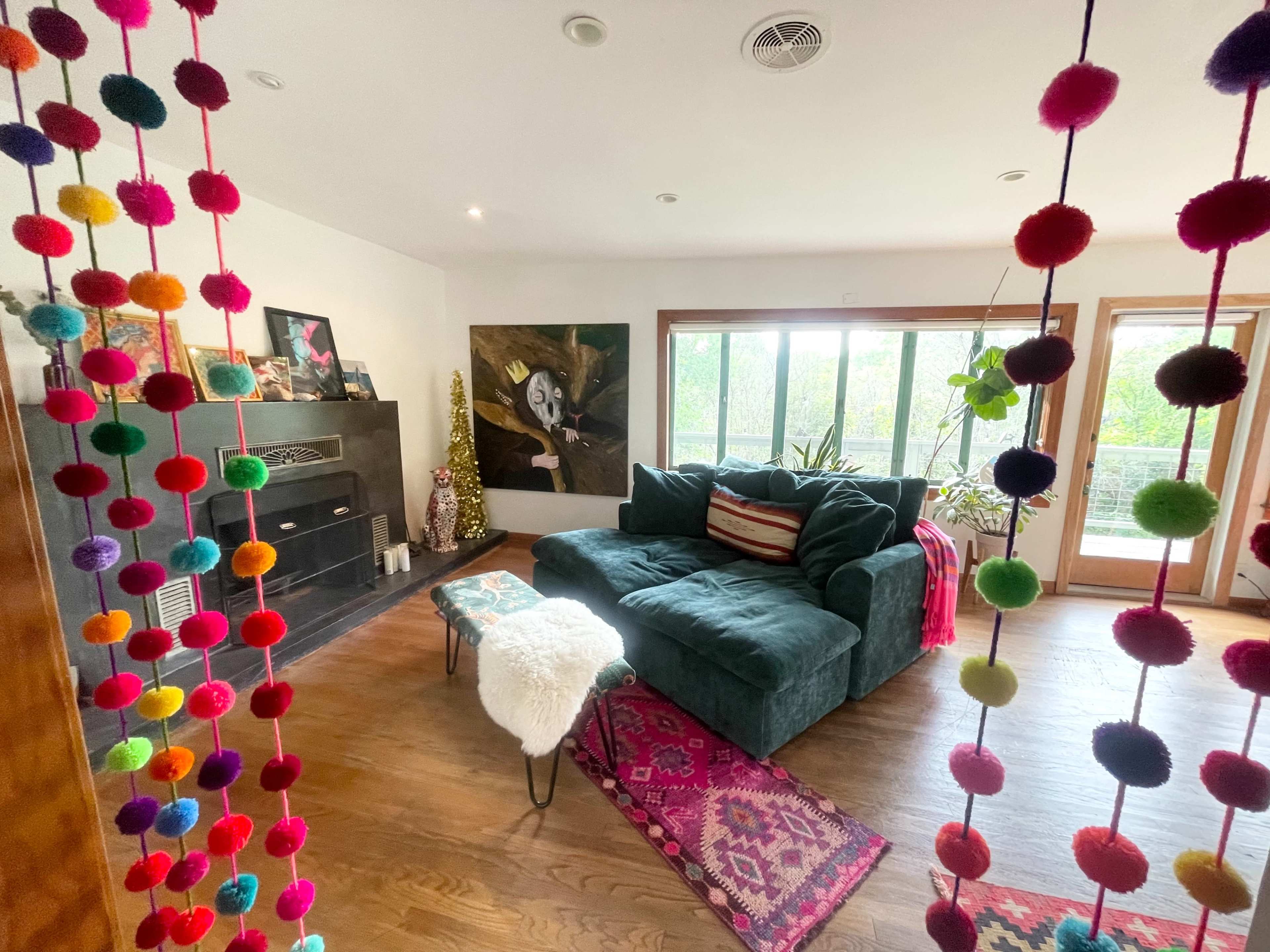 A living room features a dark green couch, a patterned rug, and colorful pom-pom decorations hanging from the ceiling.